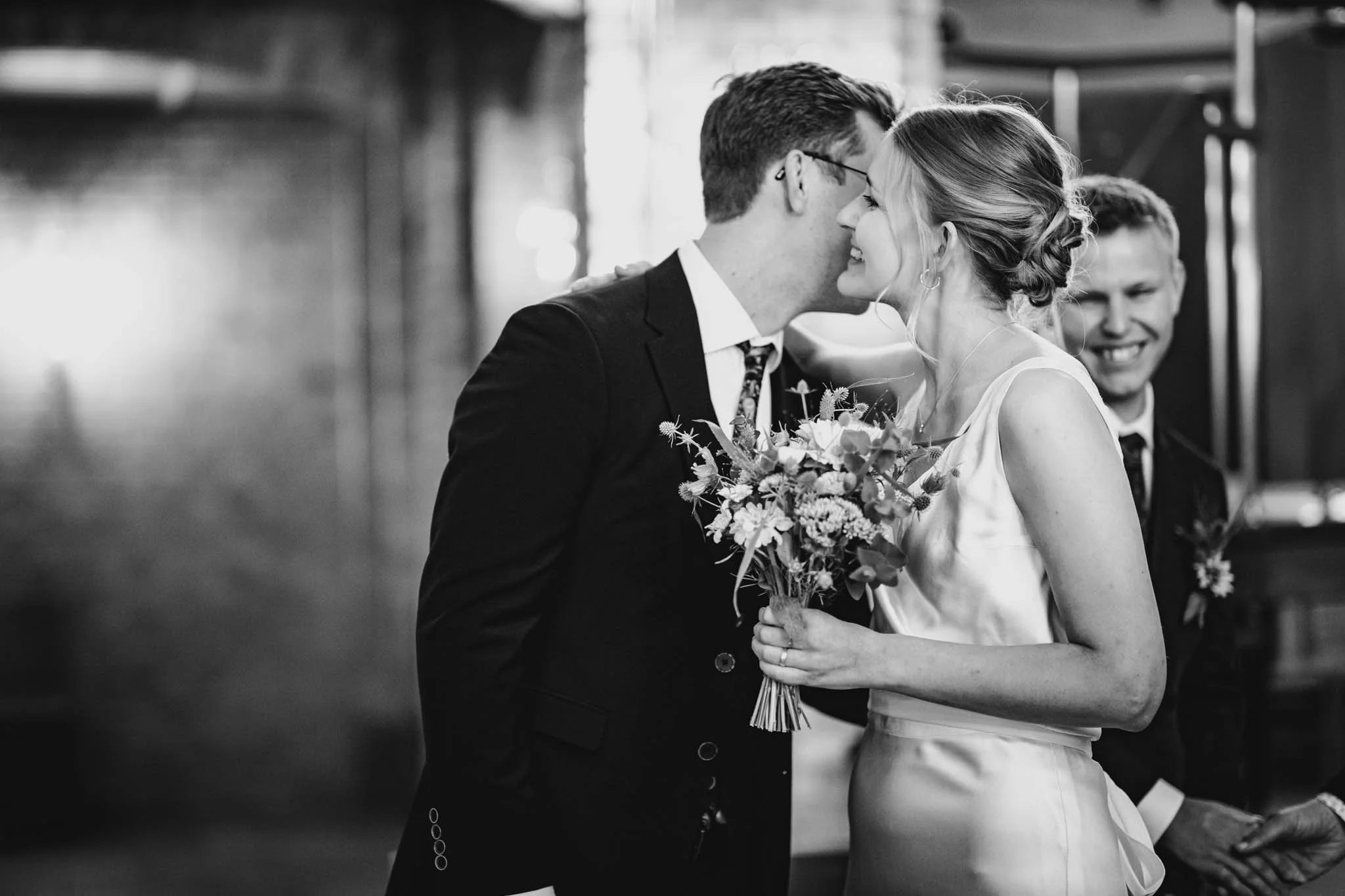 To bryllupsgæster, en mand og en kvinde, deler et øjeblik, mens de kysser næse ved en af deres største dage. Kvinden holder en buket blomster, og den mand er klædt formelt i jakkesæt og slips. Et andet bryllupspar kan ses i baggrunden, alle smiler.