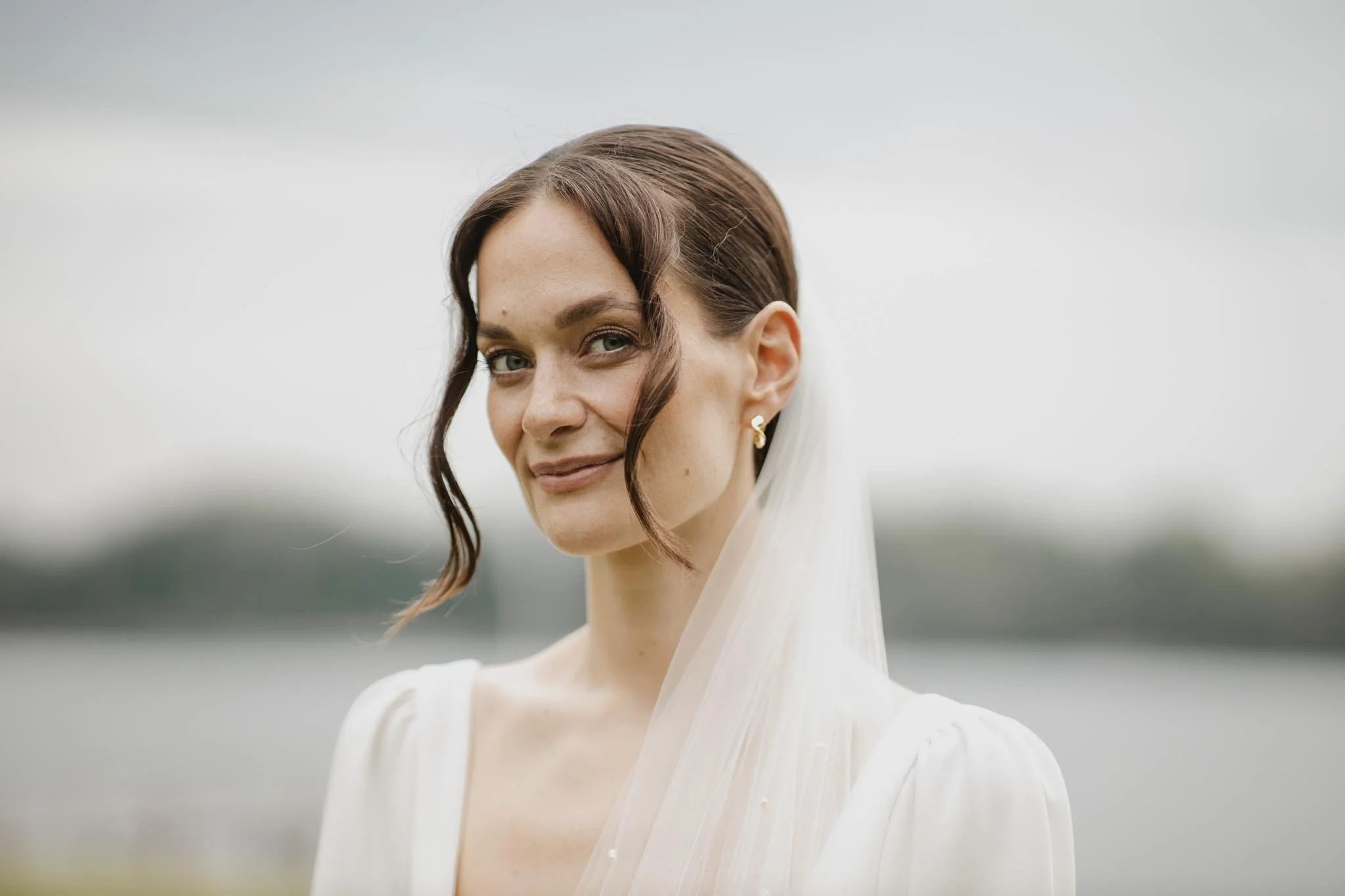 Et portræt af en glad kvinde med lyst hår og løst hår, iført en hvid kjole og øreringe, mod en neutral baggrund.