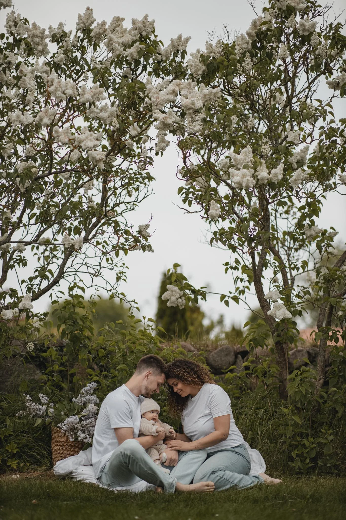 Et familieoptagelse med to voksne og et barn, siddende på jorden under blomstrende træer, med et græsk landskab i baggrunden, i et naturligt udendørs miljø.