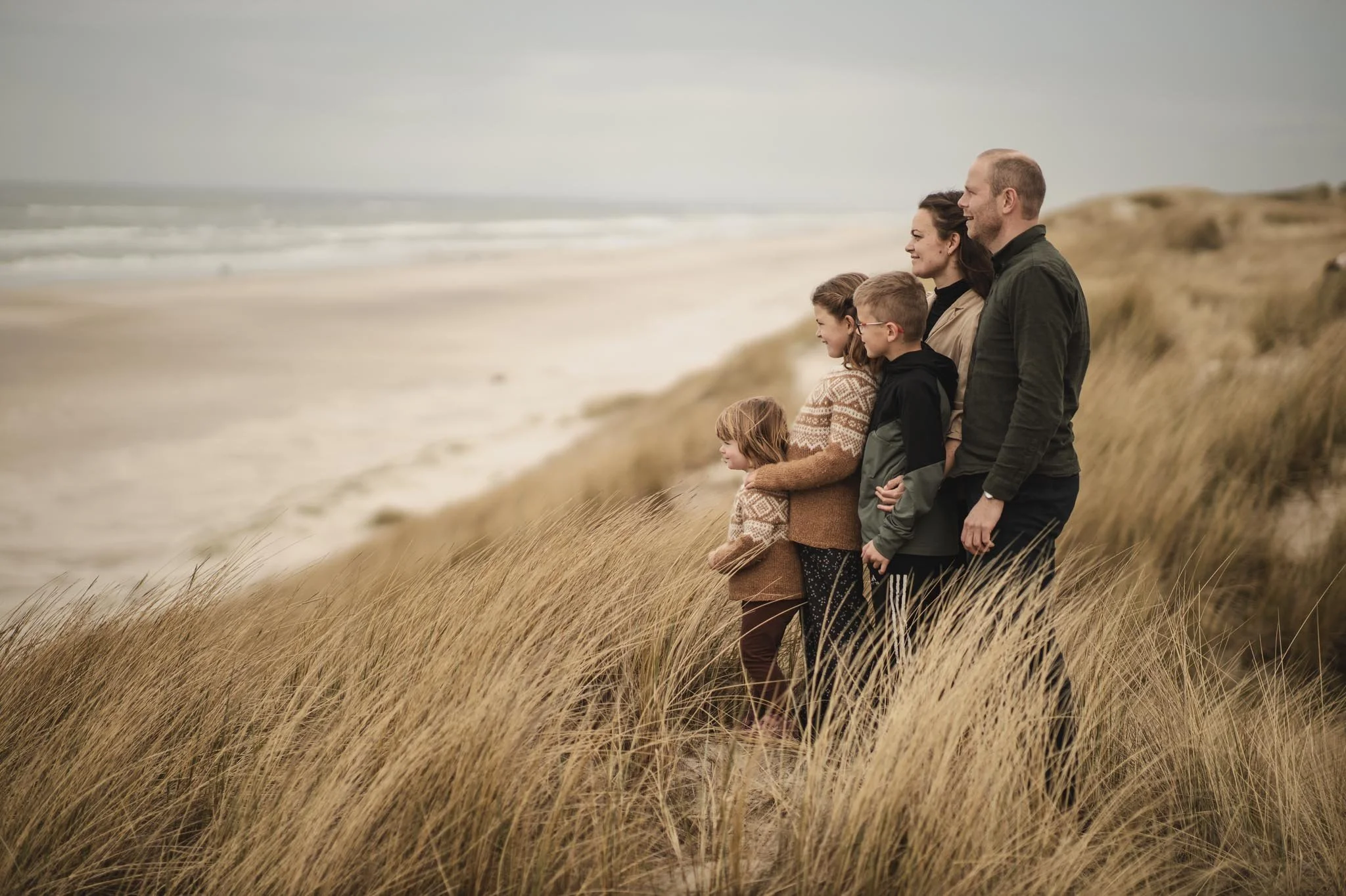 Familie står i højt græs på en klit ved stranden og nyder udsigten over havet