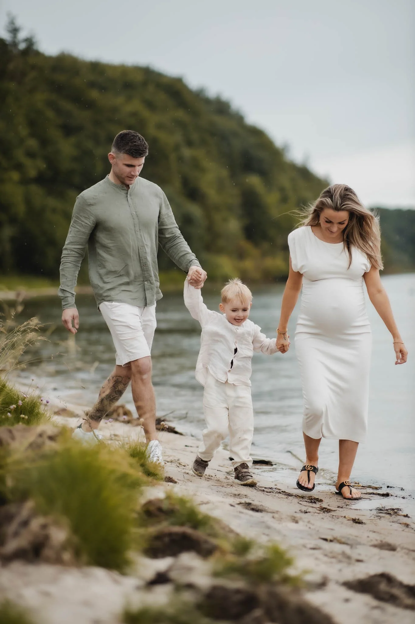 Familie går hånd i hånd ved stranden, med grønne træer i baggrunden.