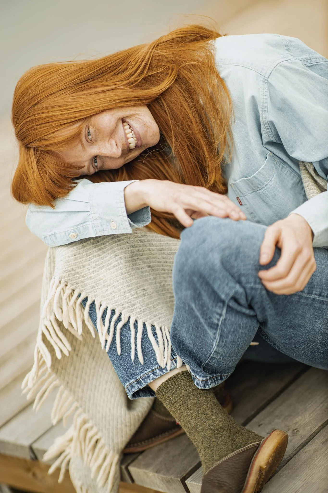 Nærbillede af en rødhaired kvinde, der smiler og ligger på en træbænk, iført en lys blå denimjakke, beige tørklæde og brune sko.