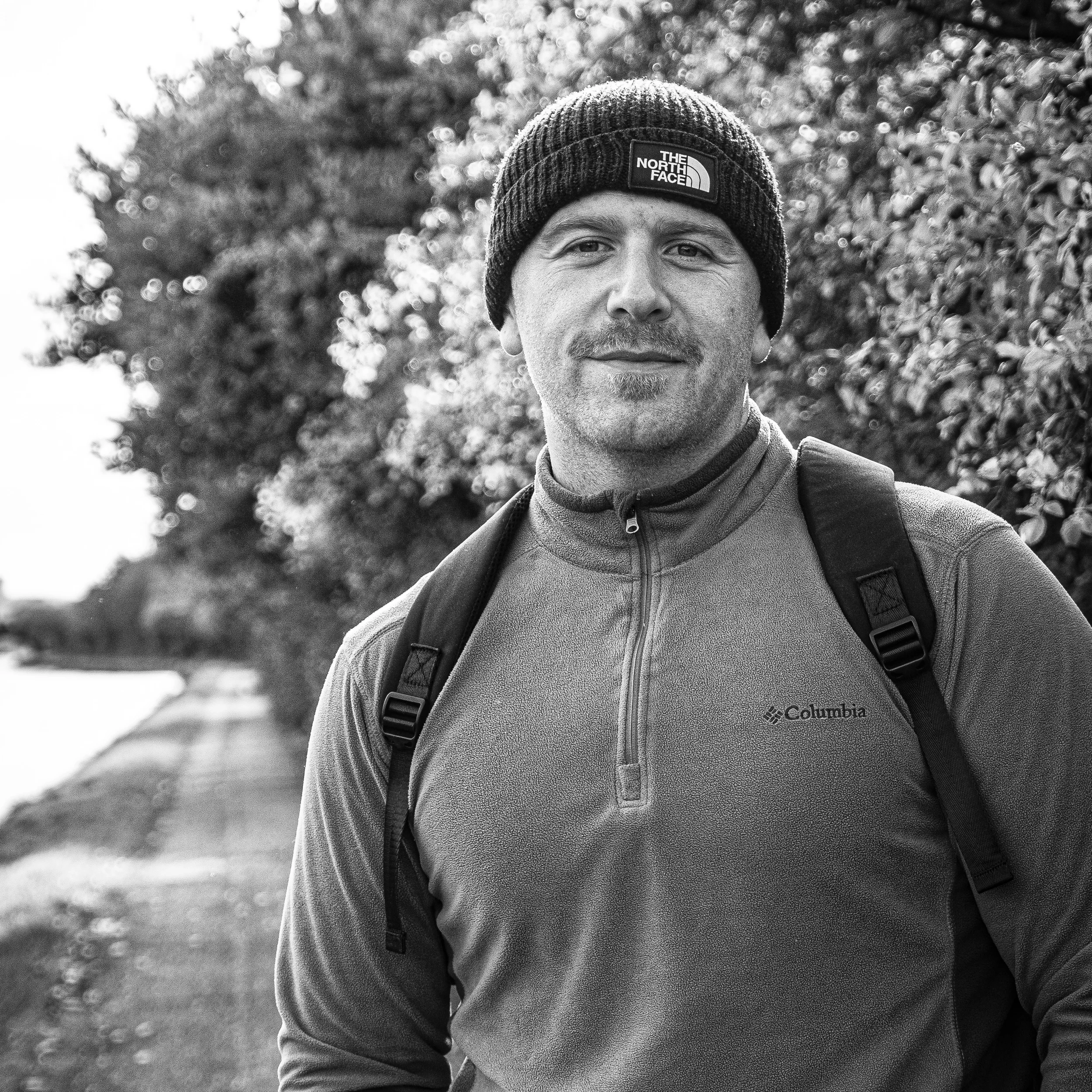 A black-and-white photo of a man wearing a beanie hat and a Columbia jacket with a backpack, standing outdoors in front of a blurred background of trees and foliage.