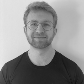 Sort-hvid portrætfoto af en mand med briller, lyst hår og skæg, iført sortT-shirt, står foran en lyse baggrund.