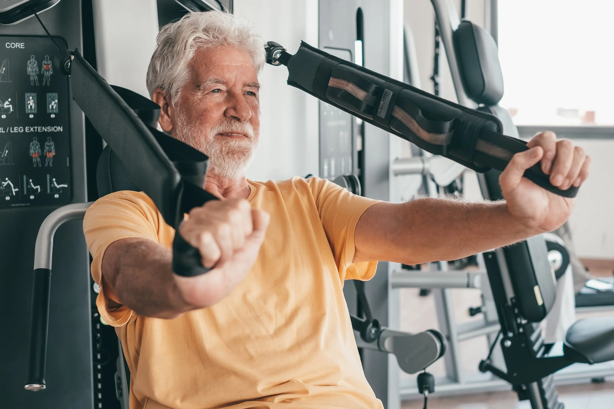 Ældre mand træner i et fitnesscenter, sidder i en maskine til overkropsstyrke, iført orange t-shirt med fokus på motion og sundhed.