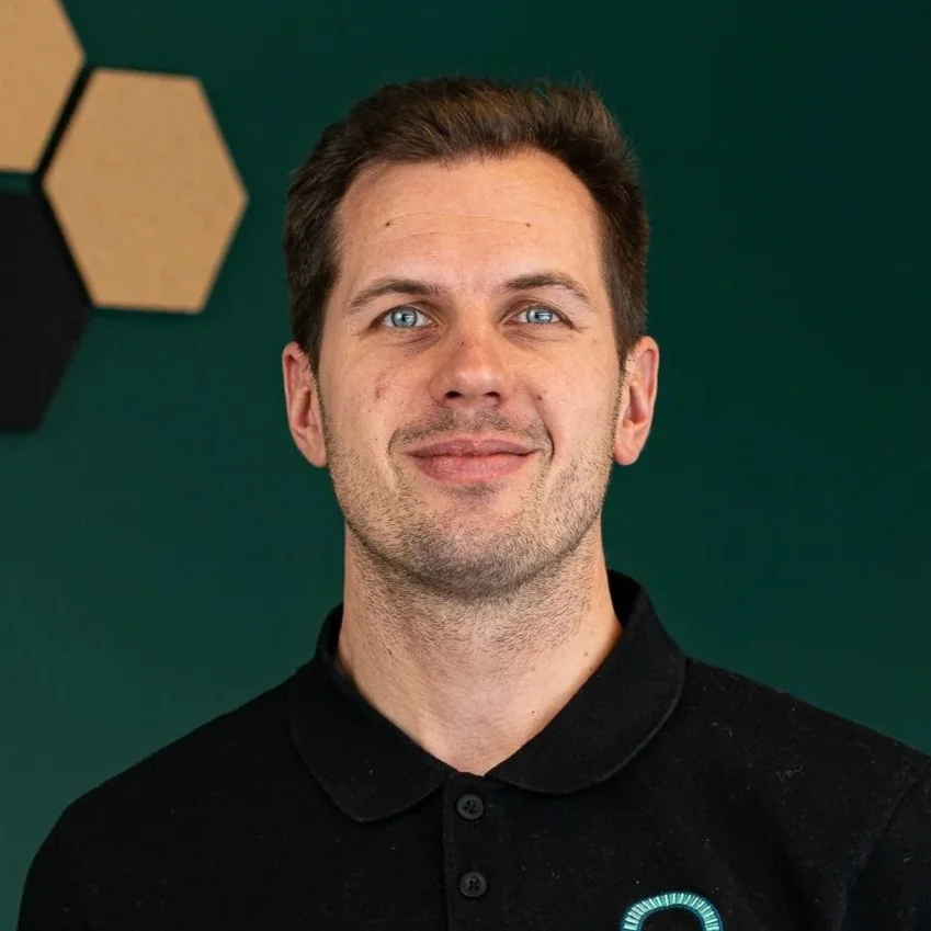 A man with short dark hair and blue eyes wearing a black polo shirt, standing against a dark green background with hexagonal wall decor.