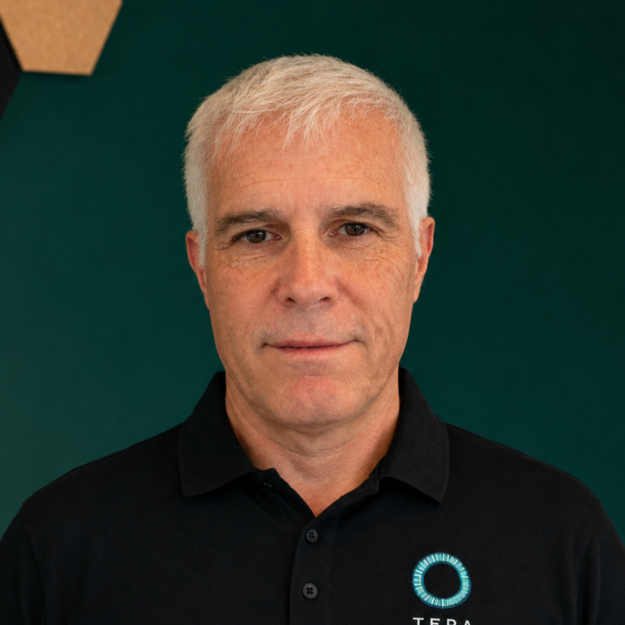 A man with short white hair wearing a black polo shirt with a logo on the chest, standing against a dark green background.