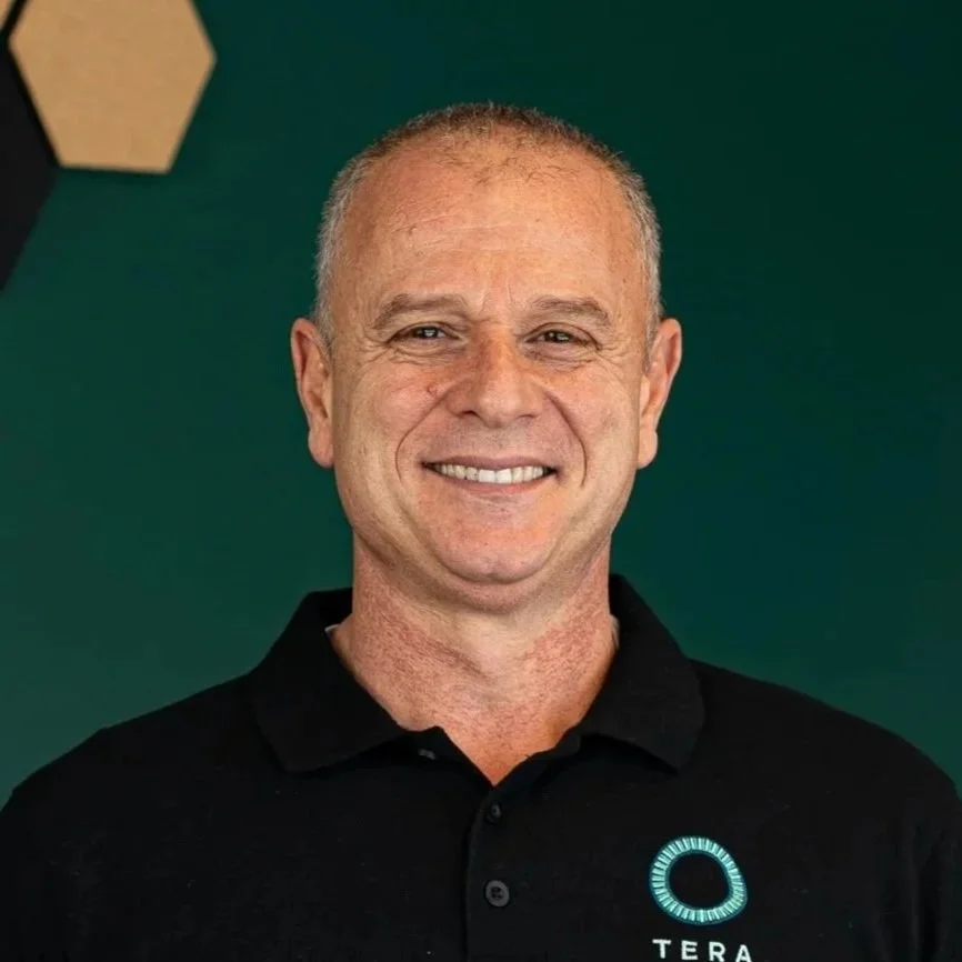 A smiling middle-aged man with short gray hair, wearing a black polo shirt with a glowing teal logo and the word "TERA" on it, standing against a dark green background.