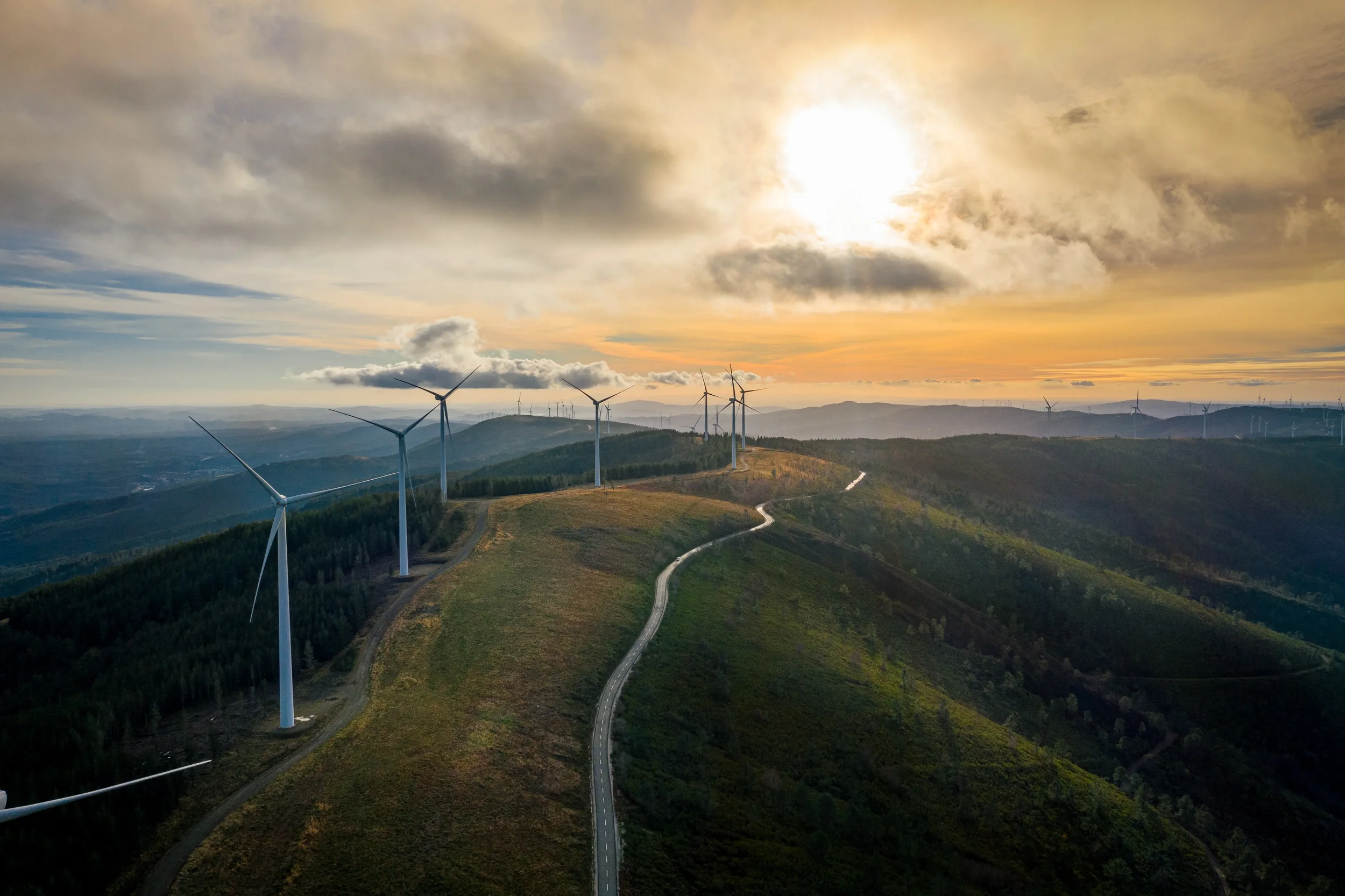 A landscape view of rolling hills with wind turbines and a winding road, under a partly cloudy sky at sunset.