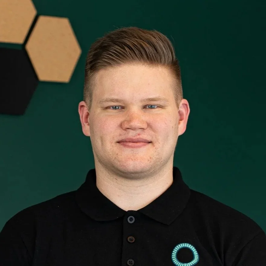A young man with short light brown hair wearing a black polo shirt with a circular logo on the chest, standing in front of a green background with hexagonal wall decorations.