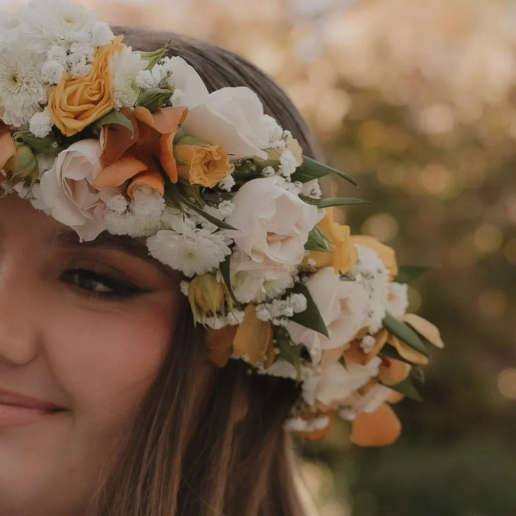 A woman wearing a floral crown made of white, yellow, and peach flowers, smiling outdoors with a blurred background of trees and sunlight.