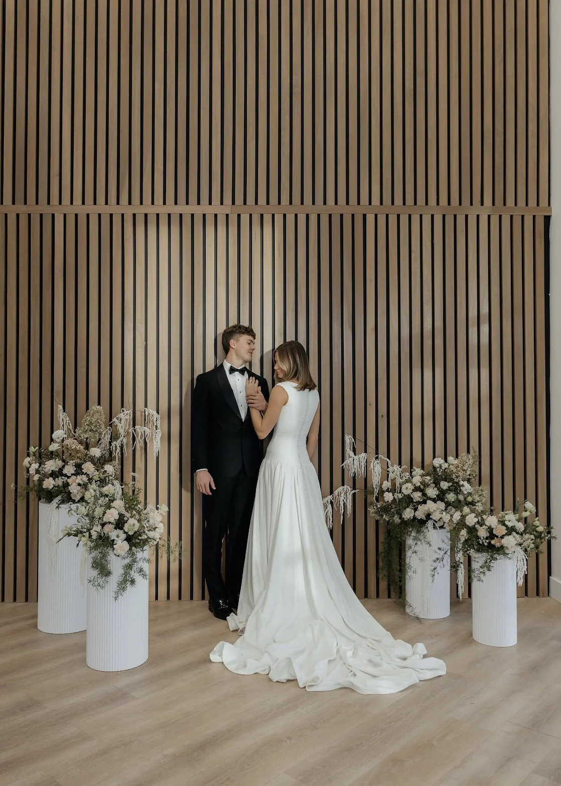 A bride and groom standing in front of a wooden slat wall, surrounded by white floral arrangements on tall white vases, during a wedding ceremony.