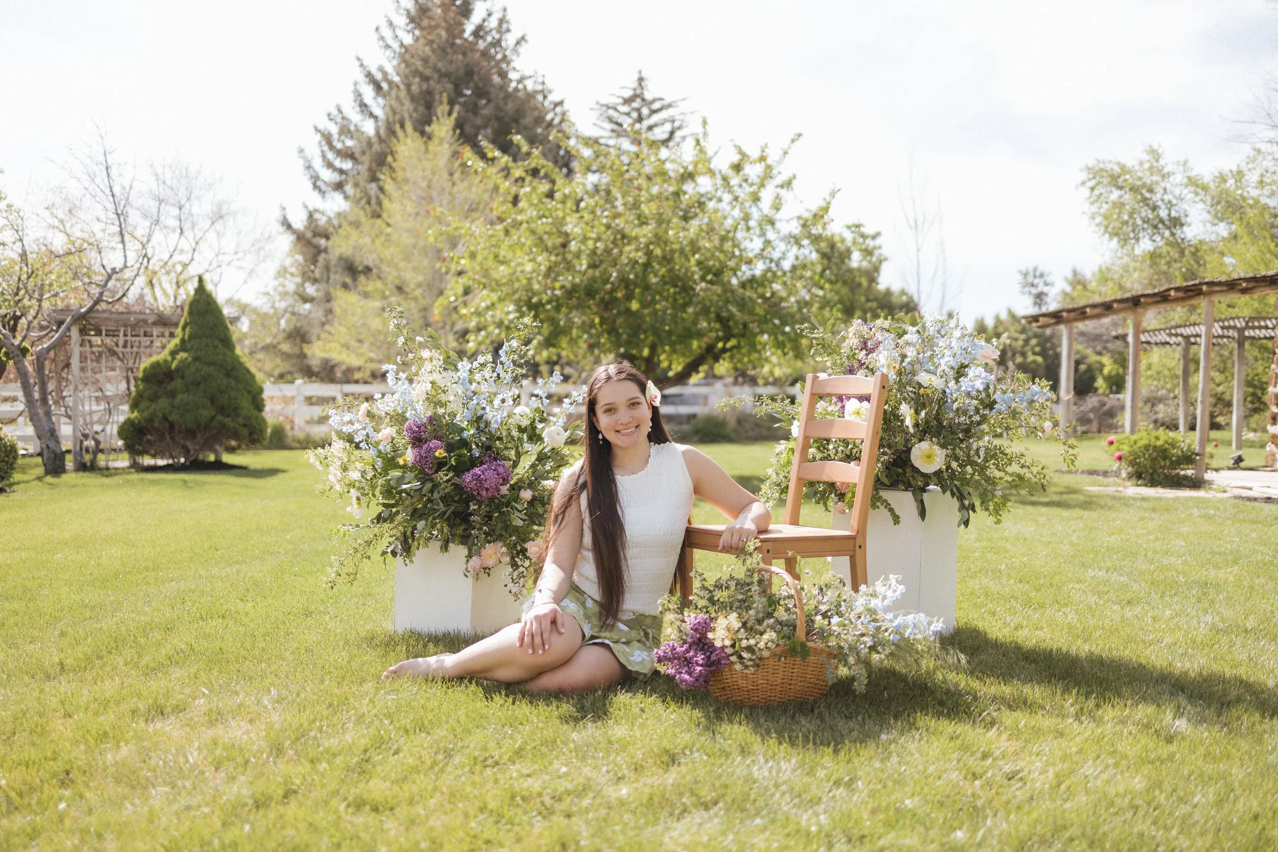 A young woman with long dark hair sitting on grass in a garden, smiling, surrounded by flower arrangements with pink, white, and purple flowers, with a wooden chair and a white wall in the background.