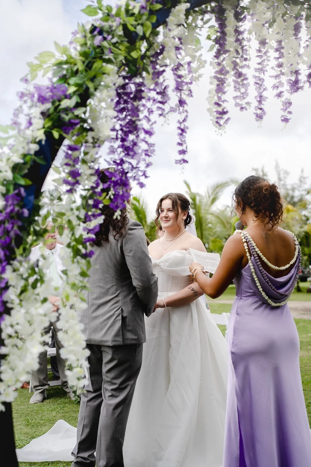 A wedding ceremony outdoors with a bride and groom exchanging vows, officiant reading from a paper, under floral decorations with purple and white flowers, greenery, and a cloudy sky in the background.