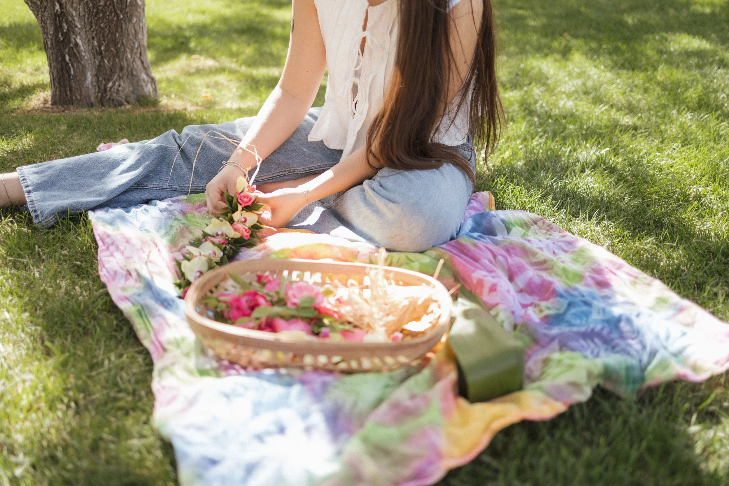 A woman sitting on a colorful tie-dye blanket on the grass, holding a small bouquet of pink and white flowers. There is a basket filled with more pink flowers in front of her, and a tree trunk is visible nearby. The scene is outdoors on a sunny day.