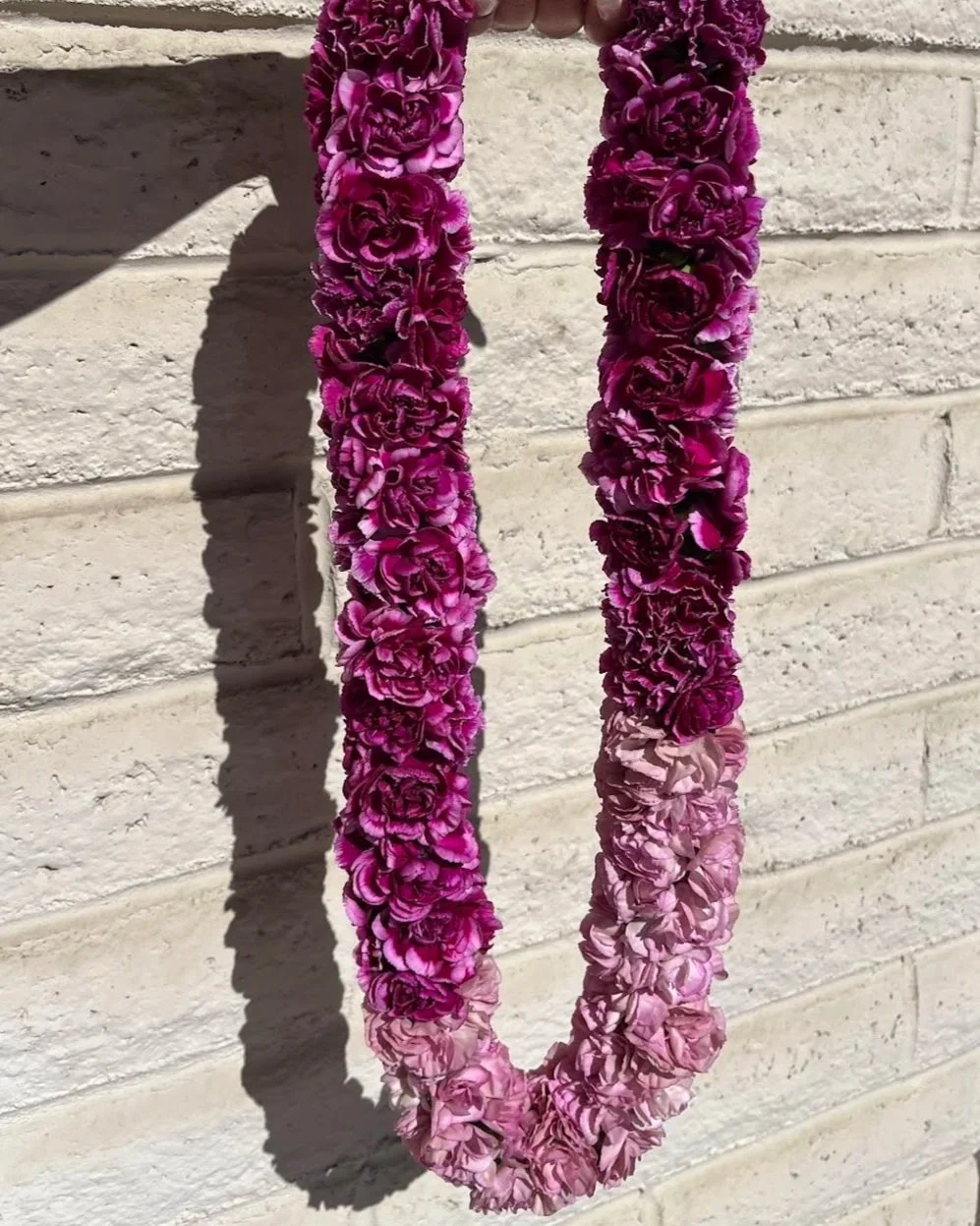 A flower lei made of purple and pink flowers hanging against a white brick wall, casting a shadow on the wall.