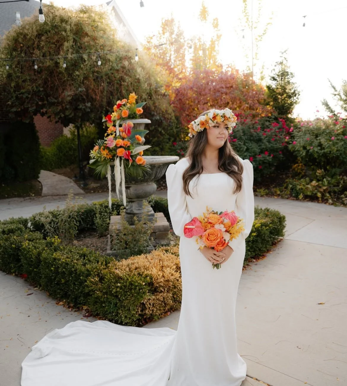 A bride in a white wedding dress holding a bouquet of orange and yellow flowers, standing outdoors with a floral crown on her head, surrounded by lush greenery and colorful flowers in the background.
