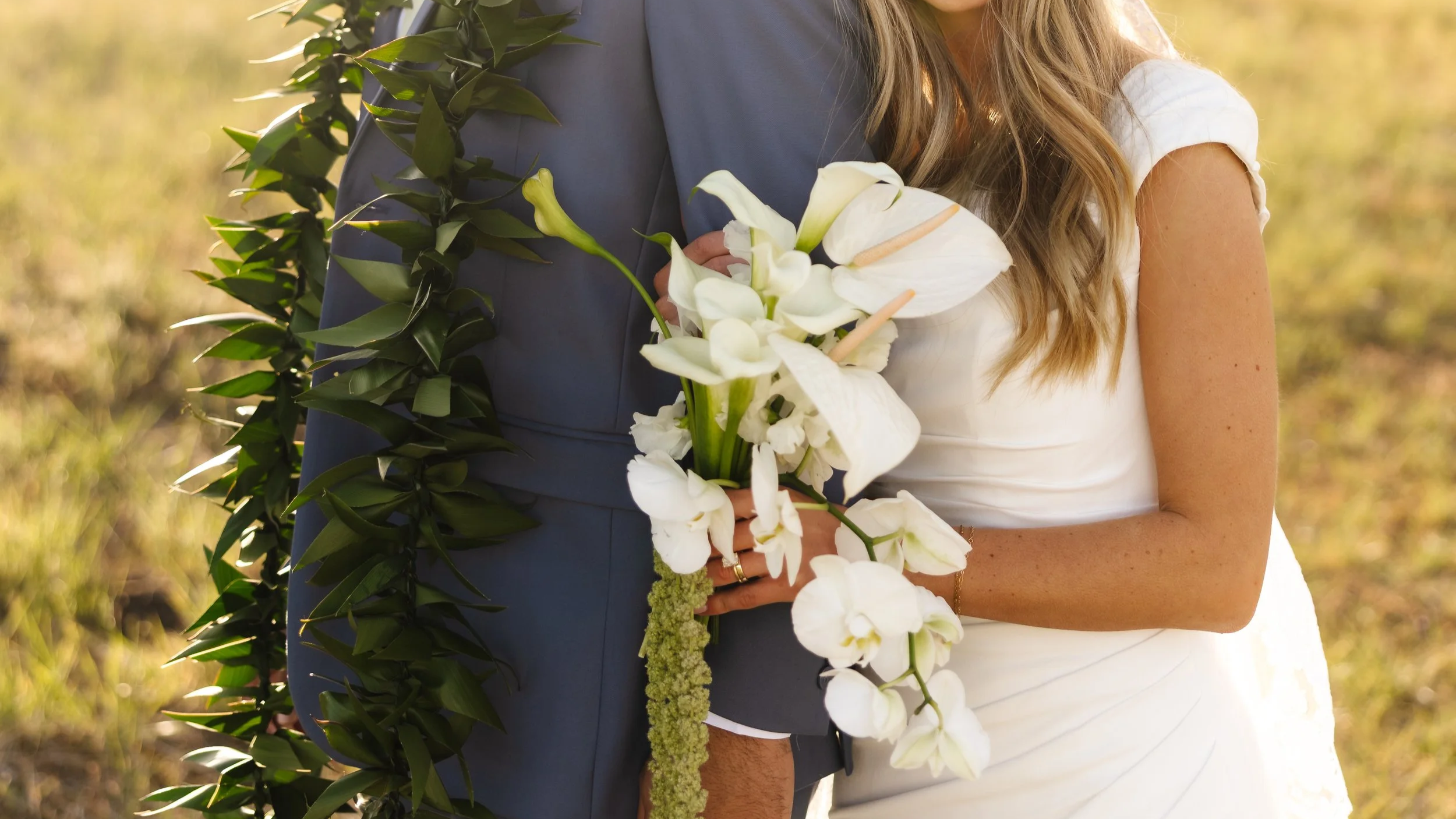 A couple dressed in wedding attire holding a bouquet of white flowers, with greenery draped over their shoulders, standing outdoors during the daytime.