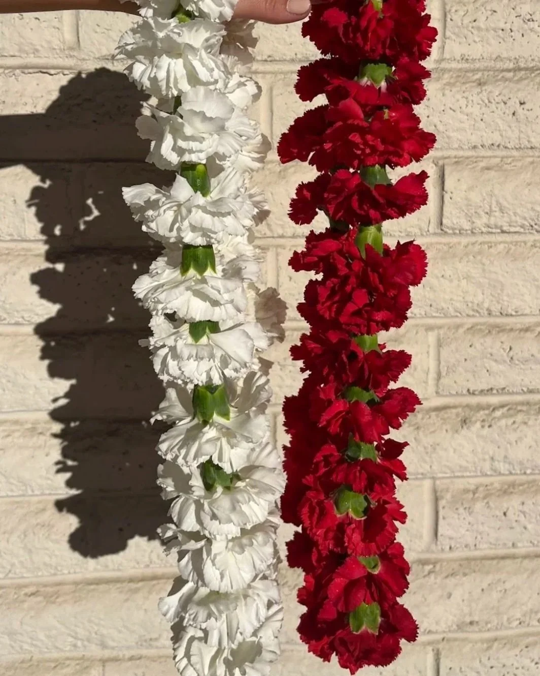 Two floral garlands, one white and one red, hanging against a beige brick wall. The shadows of the garlands are cast on the wall.