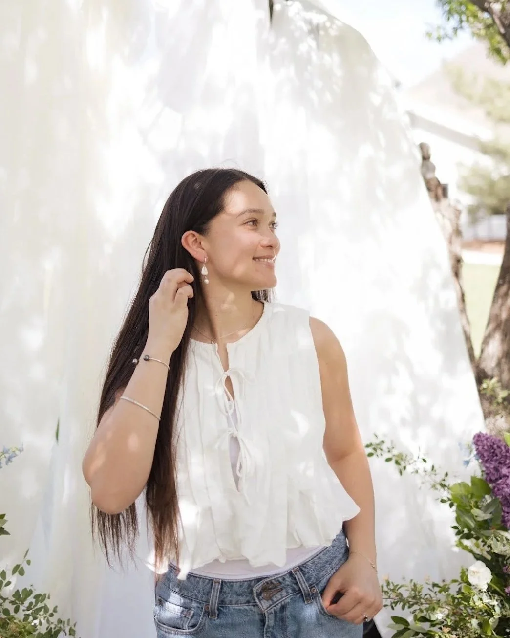 A young woman with long dark hair smiling and touching her ear with her right hand, wearing a white sleeveless top and denim shorts, in an outdoor garden setting with flowers and greenery.