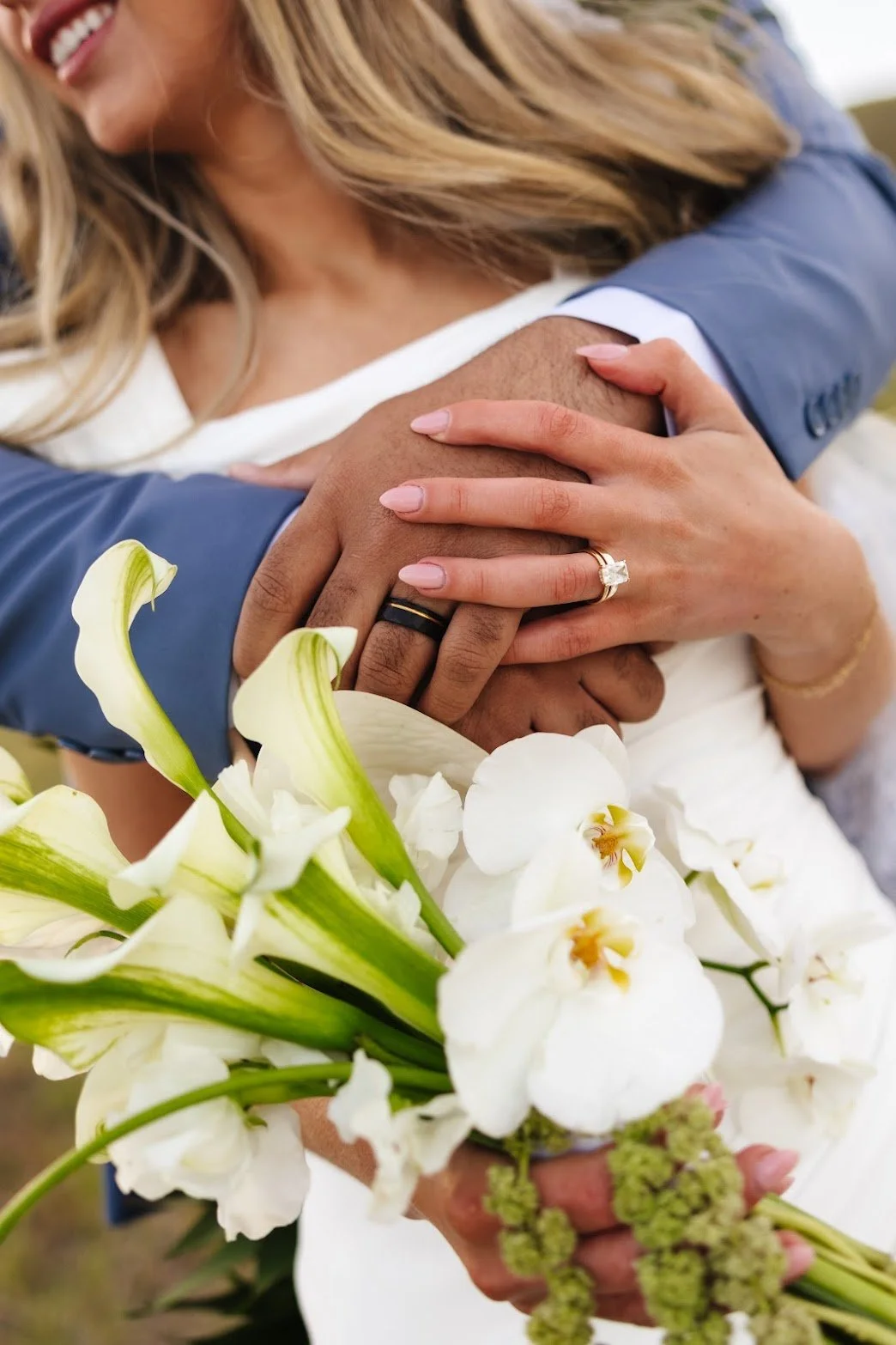Close-up of a couple's hands, with wedding rings, embracing over a bouquet of white calla lilies and orchids, with person smiling in the background.