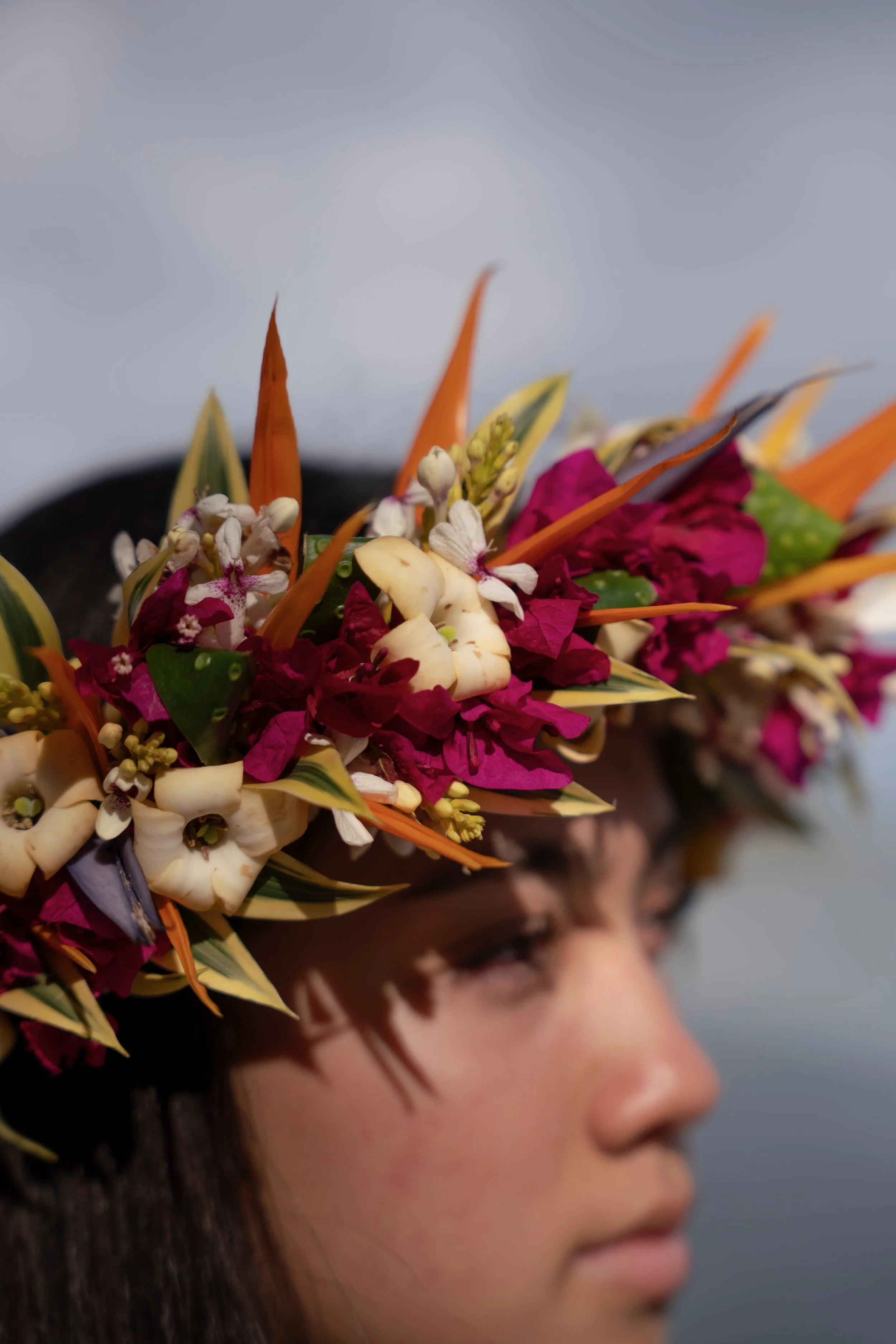 Close-up of a person wearing a colorful floral headpiece with pink, white, orange, and green flowers and leaves, with a neutral background.