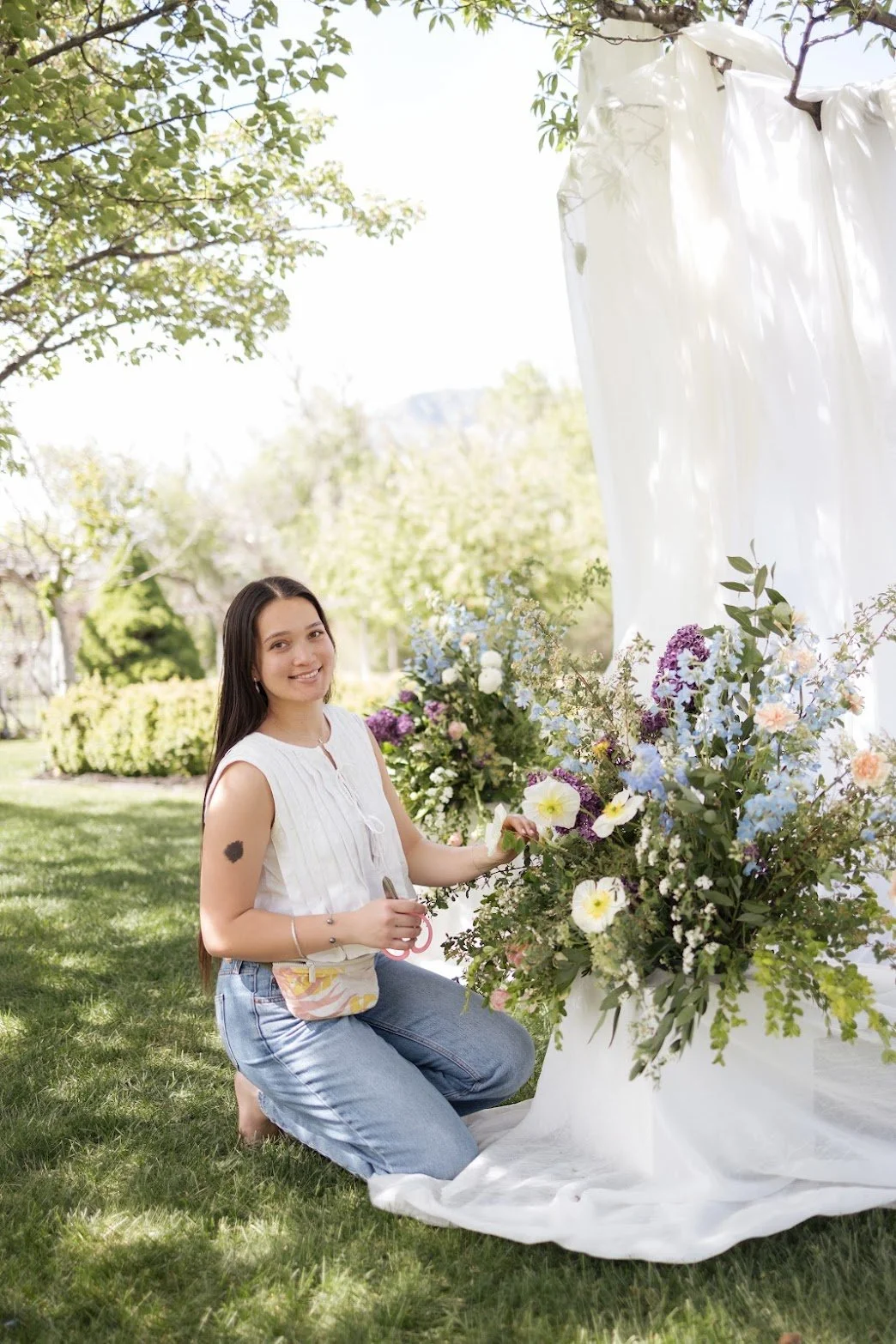 A woman kneeling outdoors on the grass, arranging a large floral display with colorful flowers and greenery, near white fabric backdrop, in bright daylight.