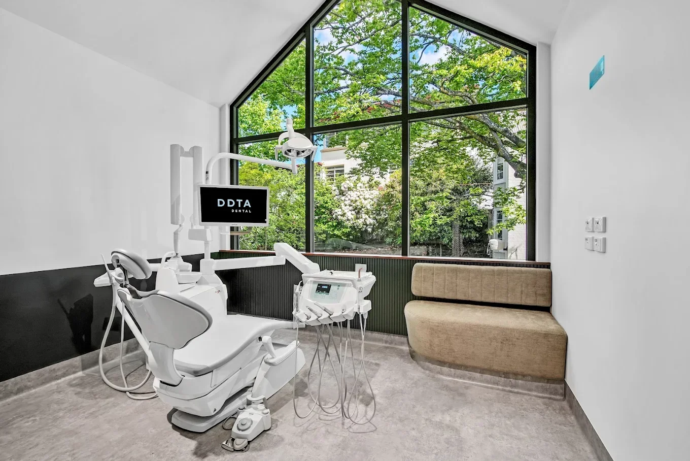 Modern dental examination room with a dental chair, medical equipment, and a large window showing green trees outside.