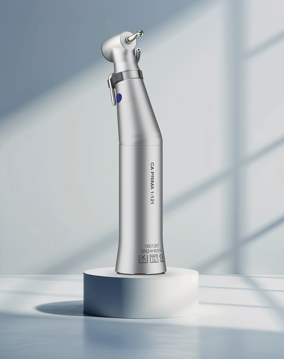 Silver dental or medical handpiece or instrument standing upright on a white circular platform, with a plain light-colored background.