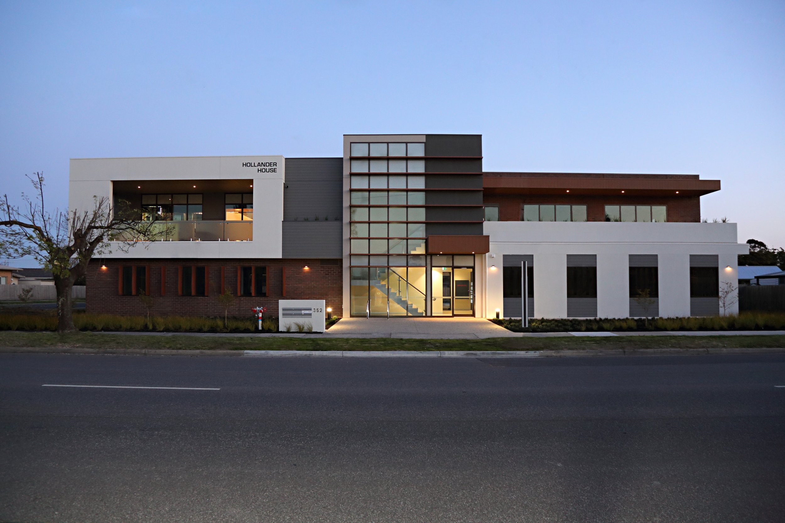 Modern multi-story residential building with white, gray, and wooden exterior finishes, large windows, and a landscaped front yard at dusk with outdoor lighting.