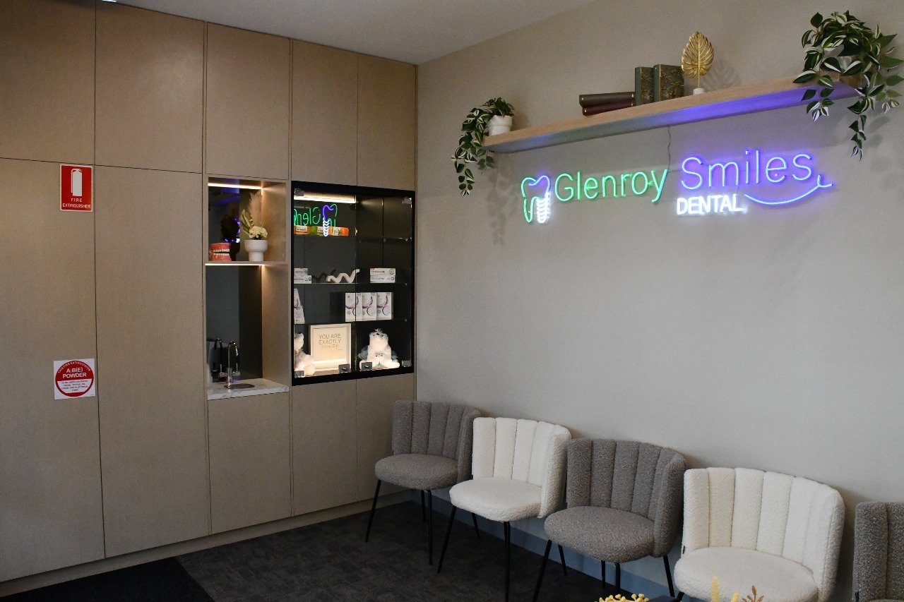 Interior of a dental office with a neon sign reading 'Glenroy Smiles Dental' on the wall, a row of mixed beige, gray, and off-white chairs, and decorative plants on shelves.