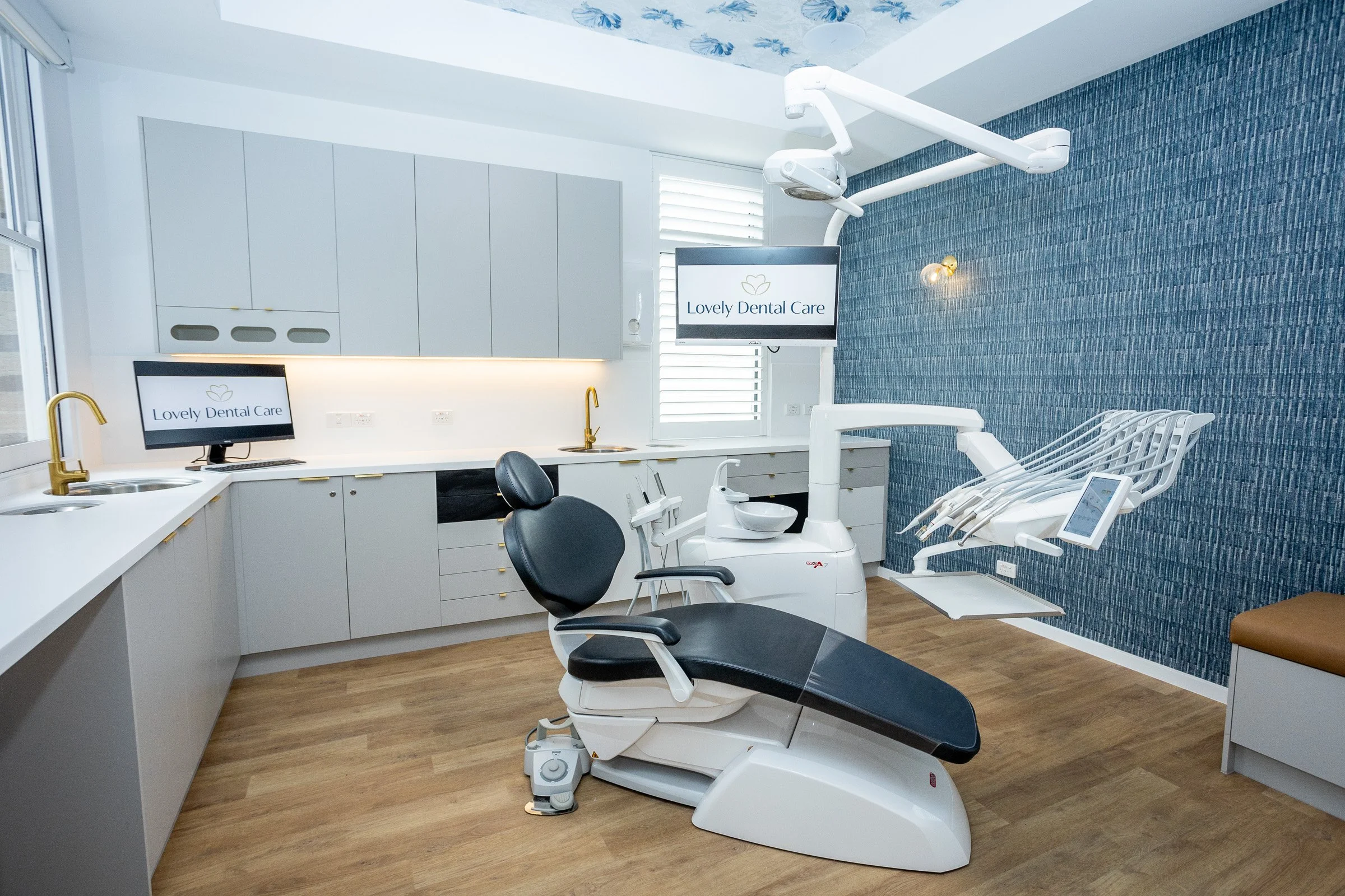 A modern dental clinic room with dental chair, monitors, and equipment, decorated in white and blue tones.