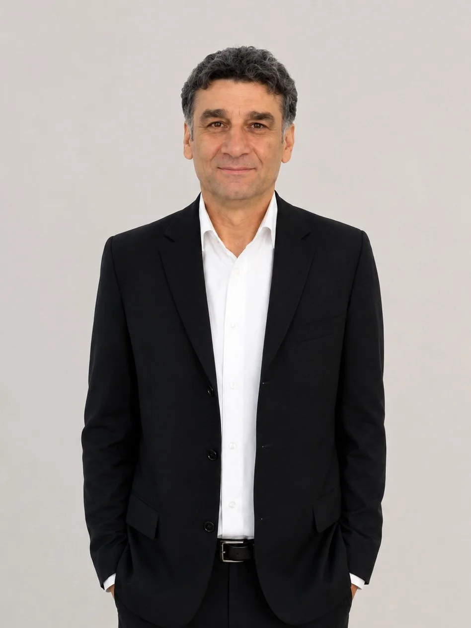 A middle-aged man with dark, curly hair wearing a black suit and white shirt standing against a plain light gray background.