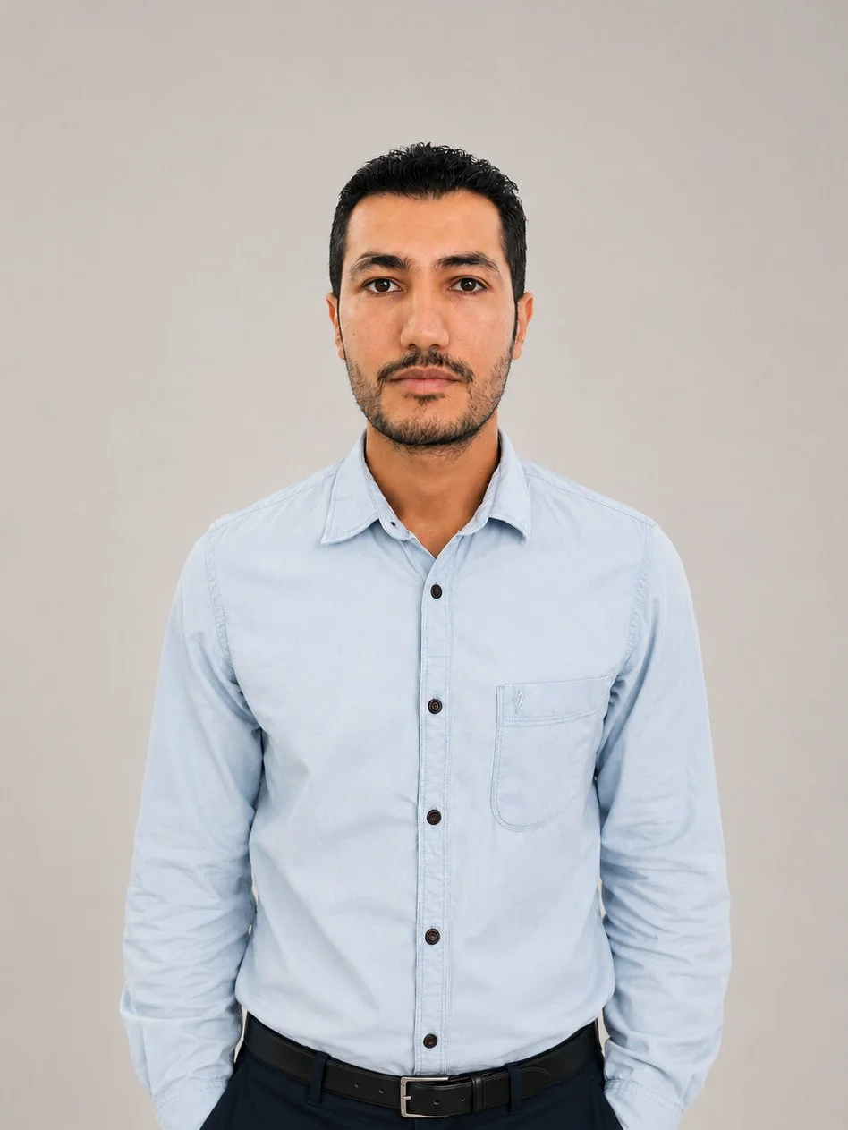 A man with dark hair and a beard in a light blue button-up shirt, standing against a plain gray background.