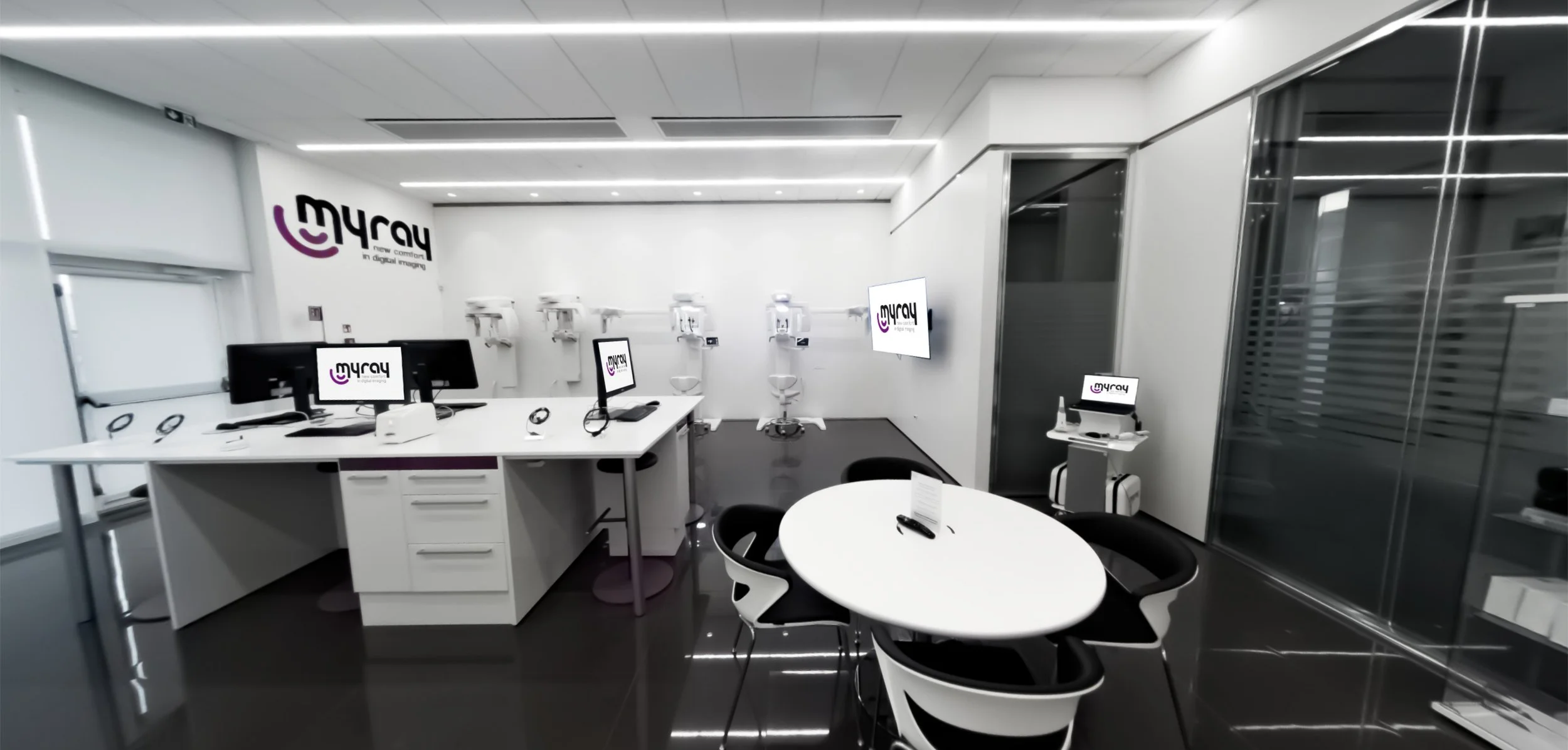 Modern hospital or clinic reception and consultation area with white walls, black flooring, and digital equipment. Multiple computer workstations with monitors displaying the 'mYray' logo, exam chairs, and medical devices aligned against the wall.