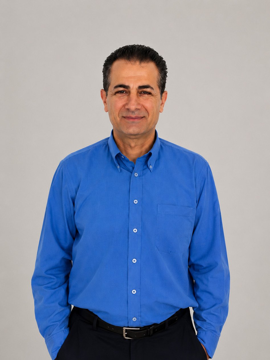 A man wearing a blue button-up shirt, standing against a plain gray background, looking at the camera with a slight smile.