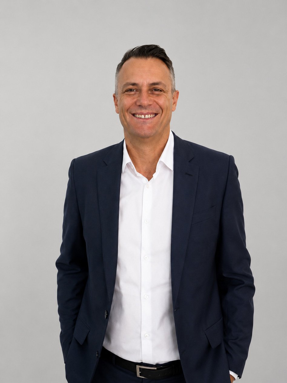 Smiling man in a navy suit and white shirt standing against a plain gray background.
