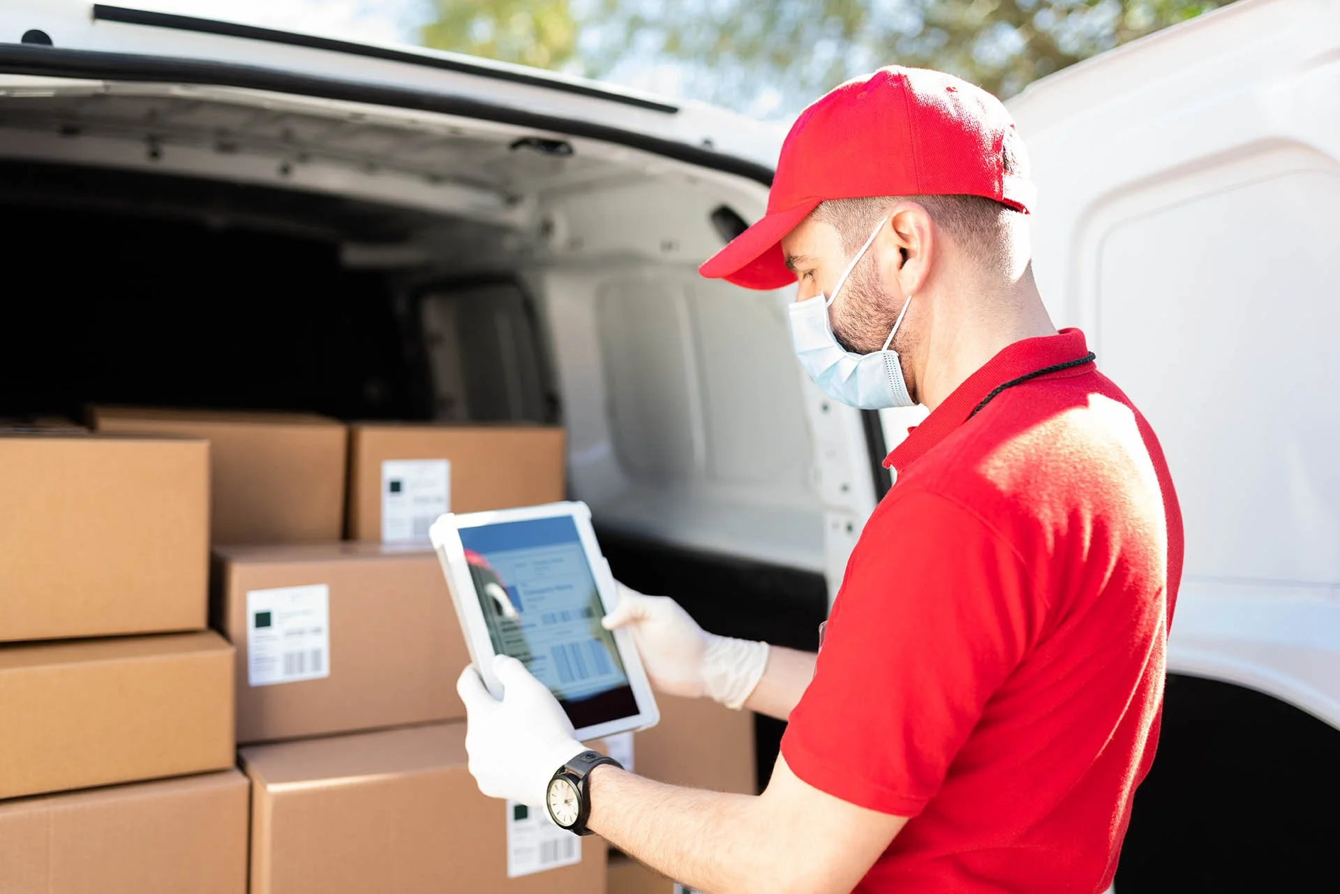 A delivery person wearing a red cap, red shirt, face mask, and white gloves standing by an open delivery van filled with boxes, using a tablet to check or scan the packages.