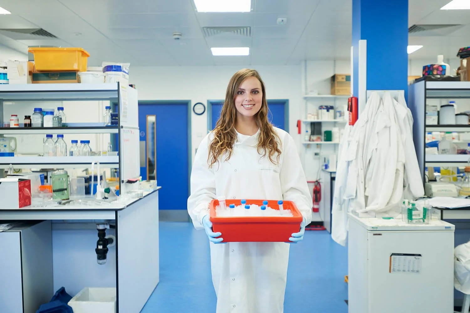 A young woman in a white lab coat and blue gloves smiling and holding a red container with test tubes in a laboratory environment.