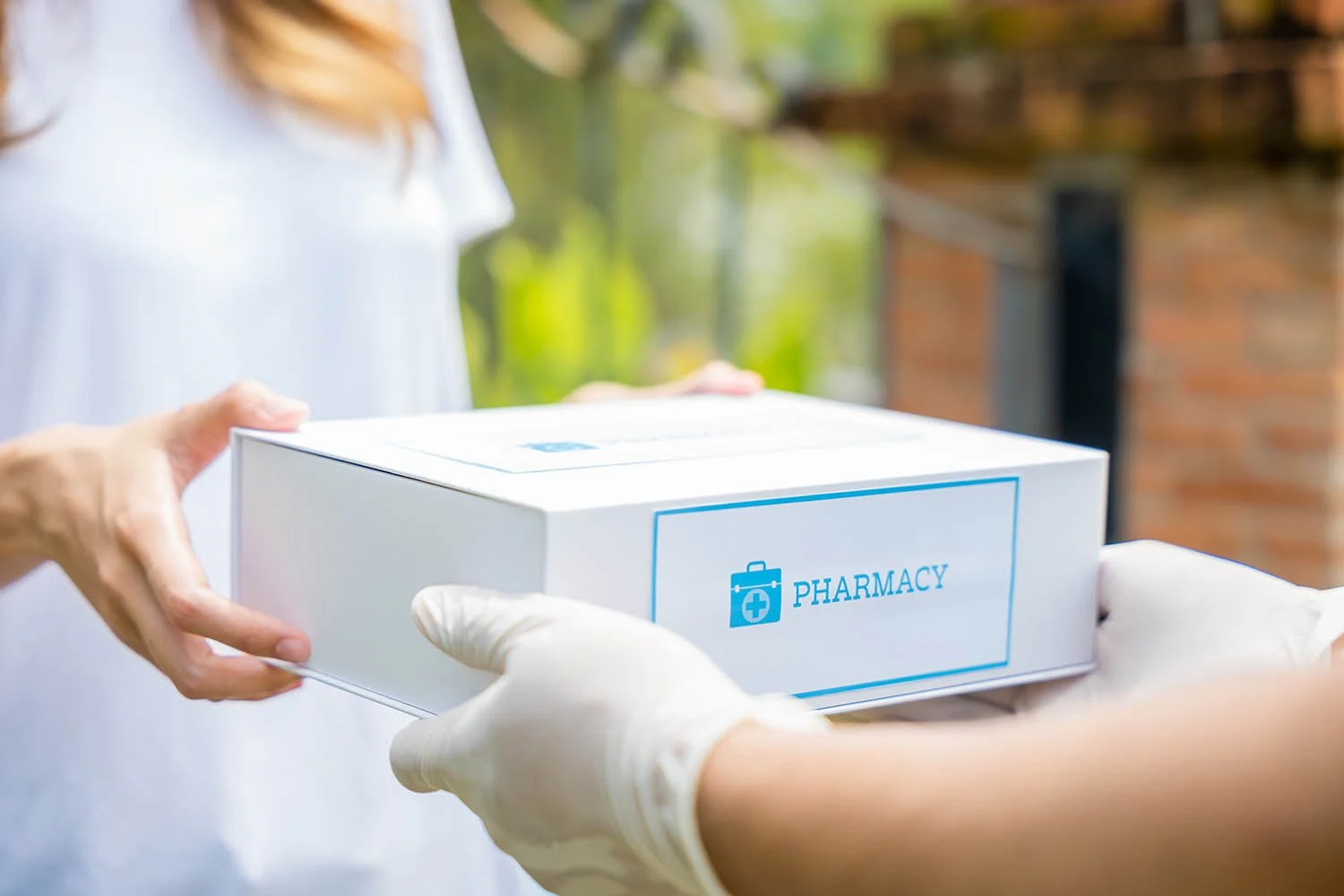 Person in white shirt receiving a pharmacy package from another person wearing gloves outdoors with a brick wall in the background.