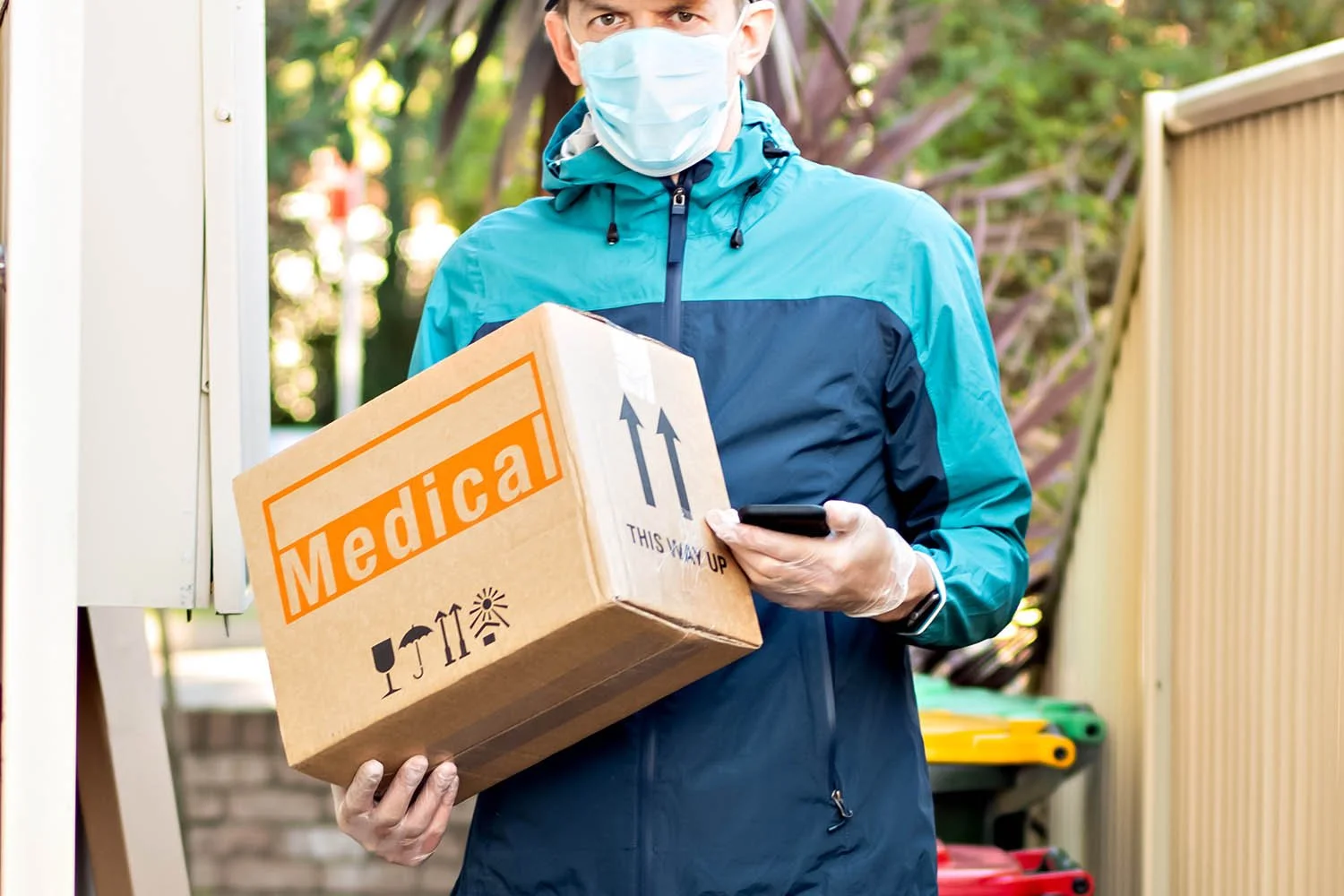 A person wearing a face mask, gloves, and a blue jacket, holding a cardboard box labeled 'Medical' with upward arrows and handling instructions, standing outdoors.
