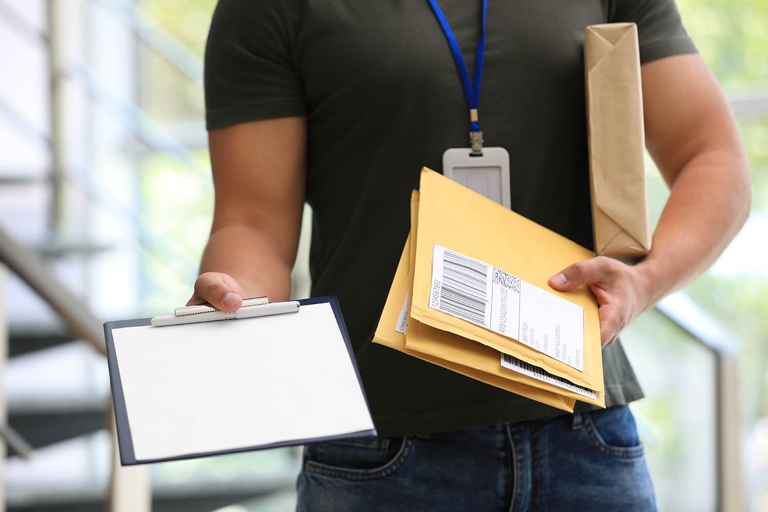Person holding a clipboard and packages, wearing a black shirt and a lanyard with an ID badge, in a warehouse or office setting.