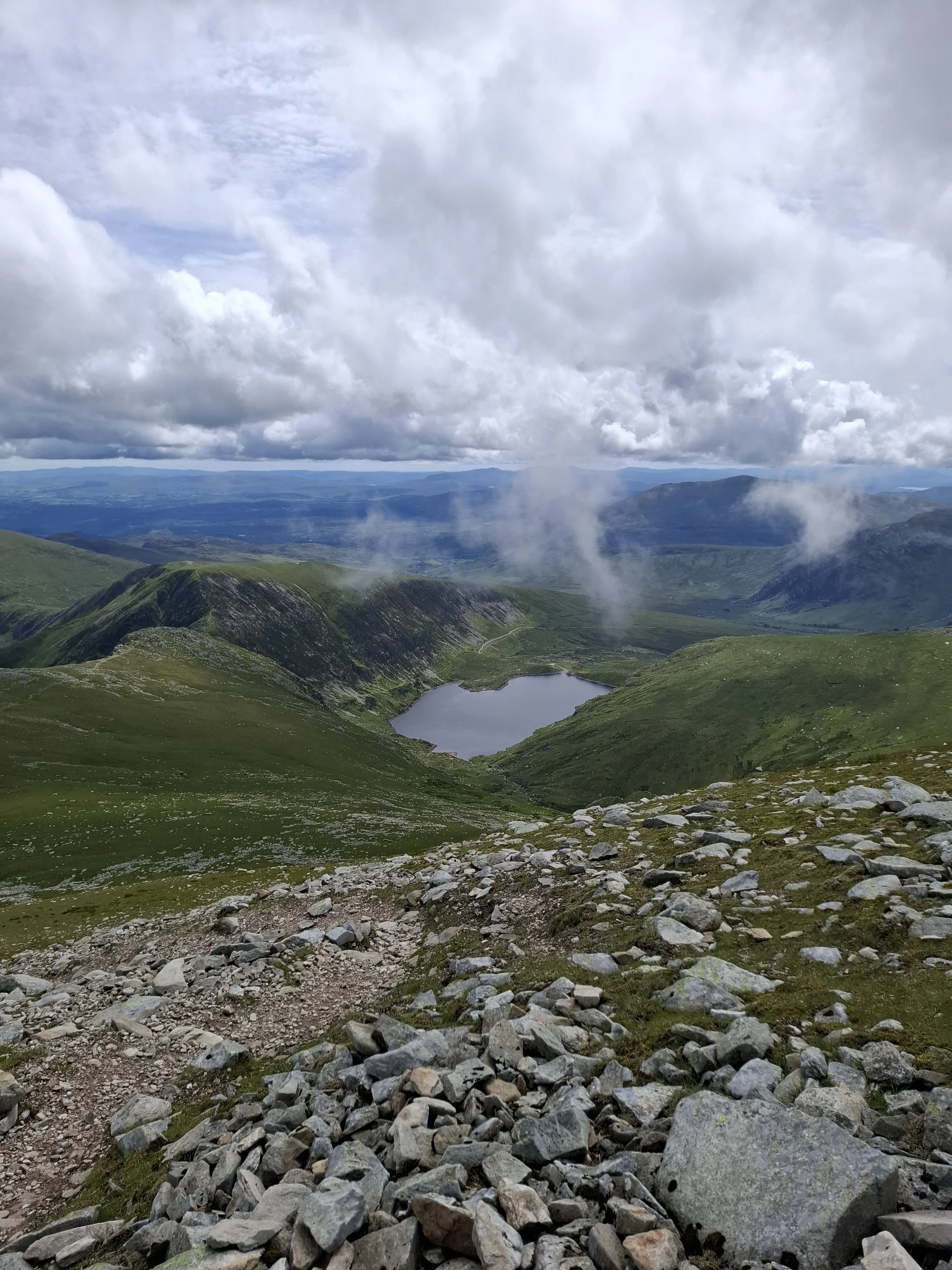 A mountainous landscape with green hills, a lake, and a cloudy sky.