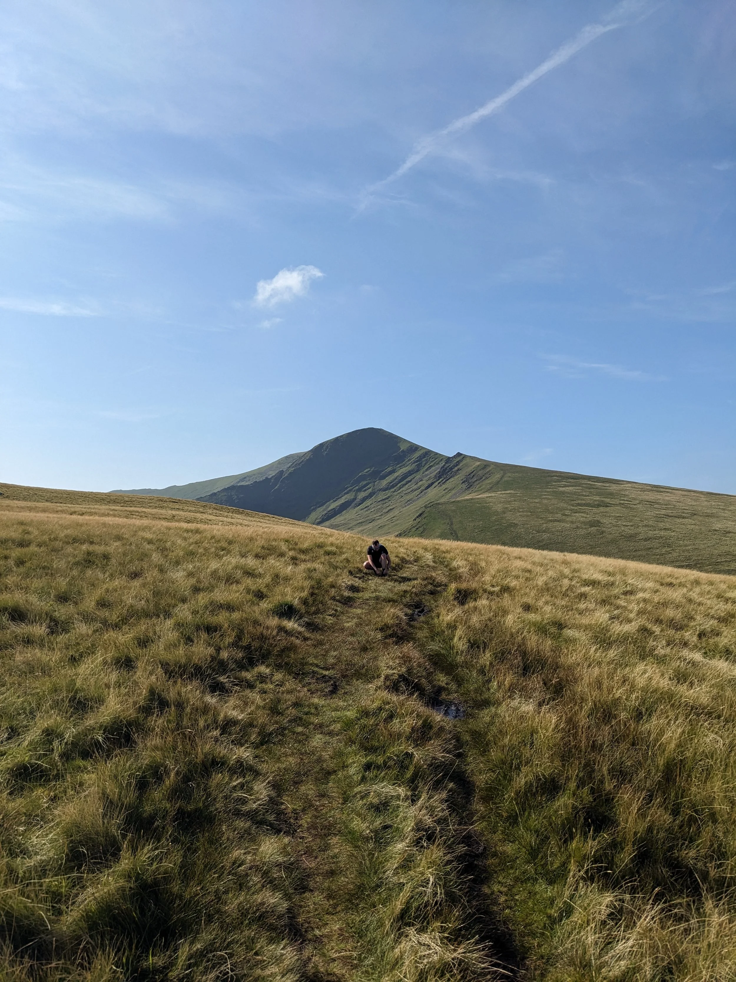 A person crouching on a grassy hillside with mountains in the background under a blue sky with a few clouds.