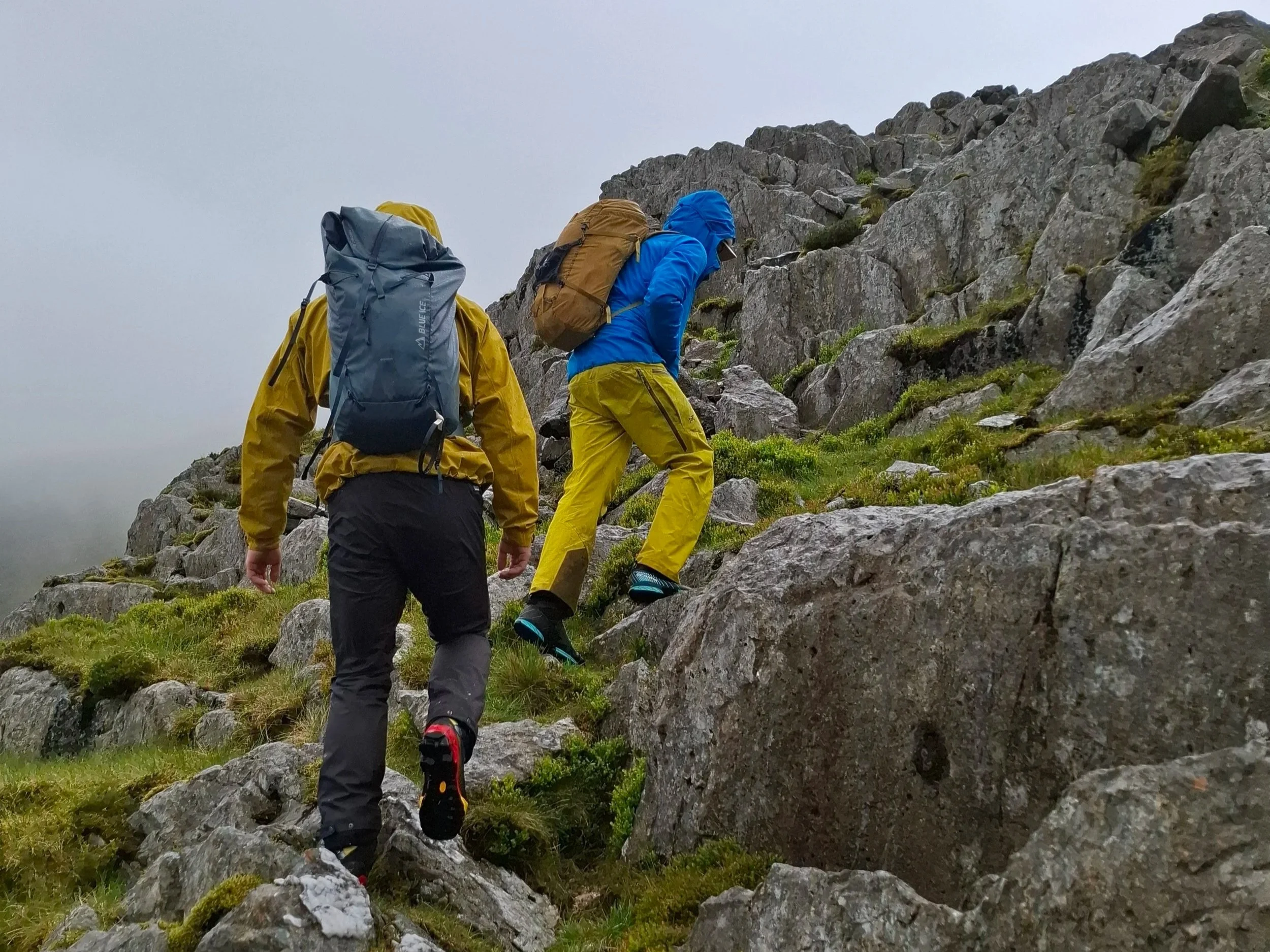 Two hikers climbing a rocky mountain trail on a foggy day, dressed in bright outdoor gear with backpacks.