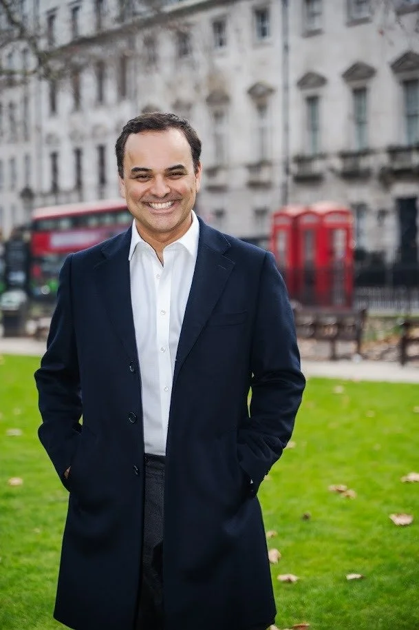 A smiling man in a dark navy coat and white shirt stands with hands in pockets on a grassy area with buildings, a red telephone booth, and a red double-decker bus in the background.