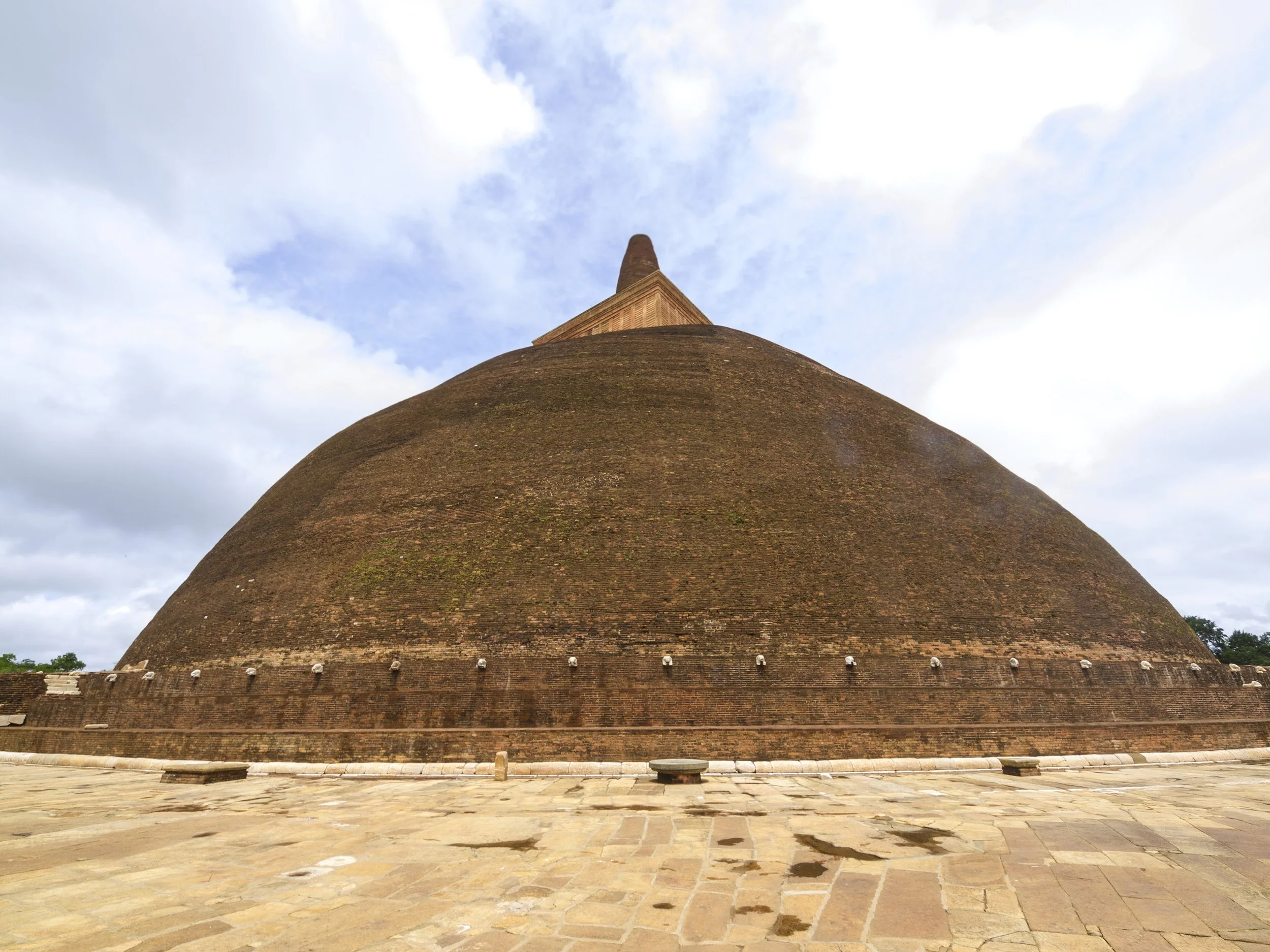 A large ancient brick stupa with a rounded bottom and a pointed top, situated on a paved open area under a partly cloudy sky.