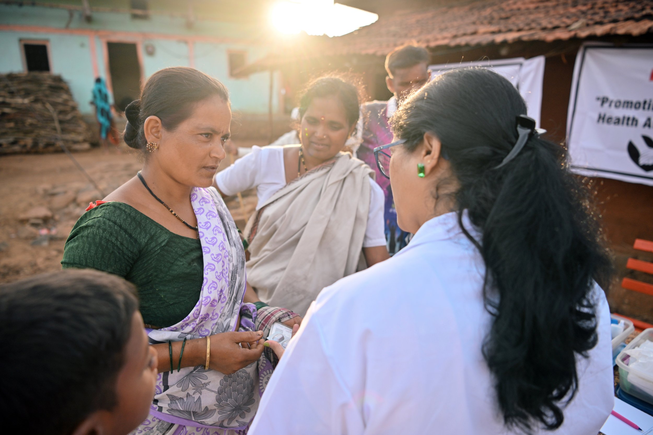 A group of women engaging in a health-related activity outdoors in the late afternoon sunlight, with a rural setting and buildings in the background.