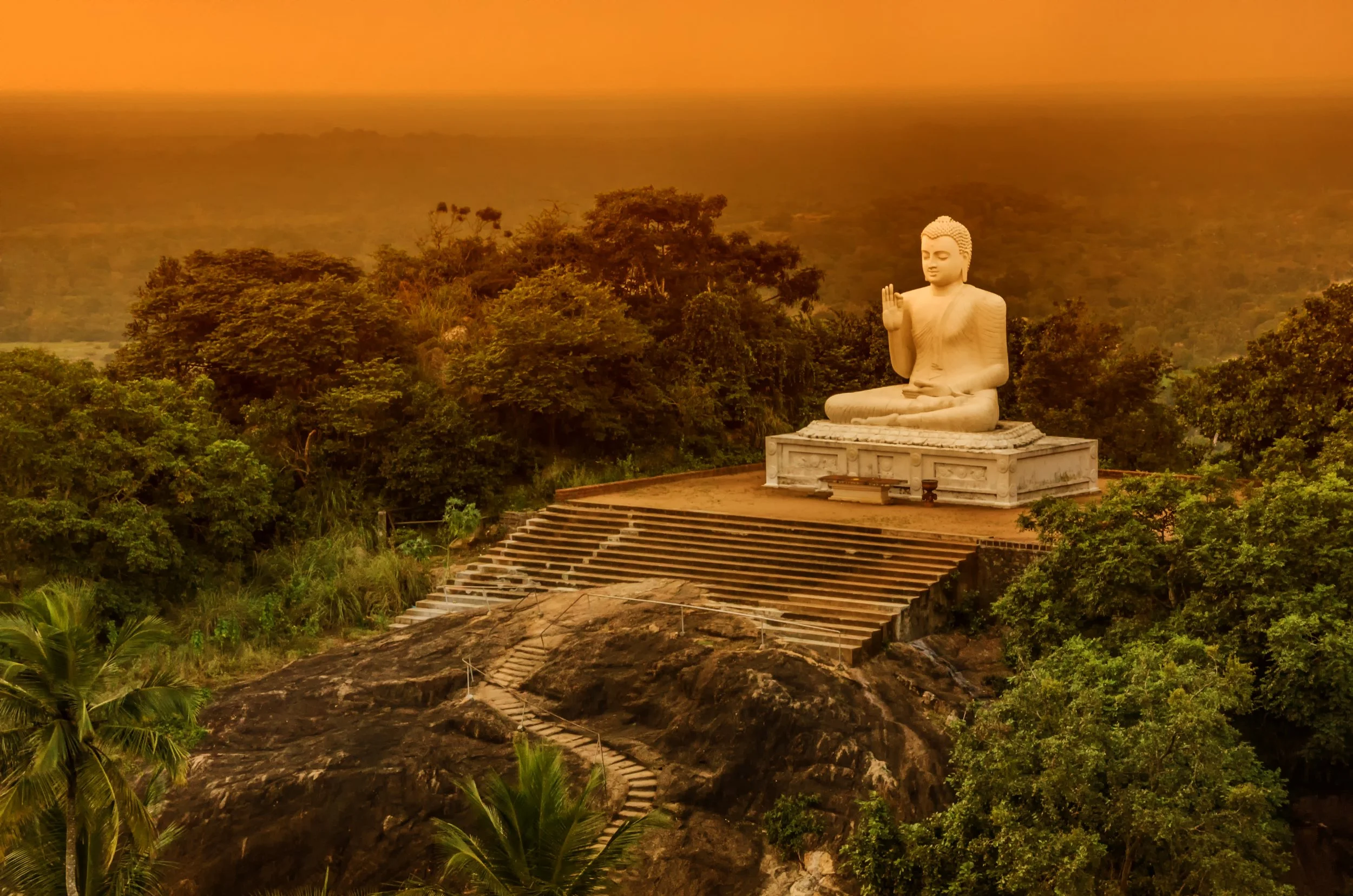 A large white Buddha statue in a seated meditation pose with one hand raised in a blessing gesture, surrounded by lush green trees and foliage, with a staircase leading up to it, under an orange evening sky.