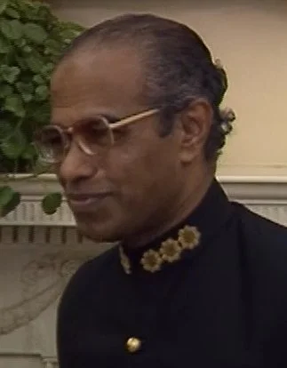 A man with glasses and curly hair is wearing traditional Indian attire with floral embroidery, standing in front of a fireplace with greenery.